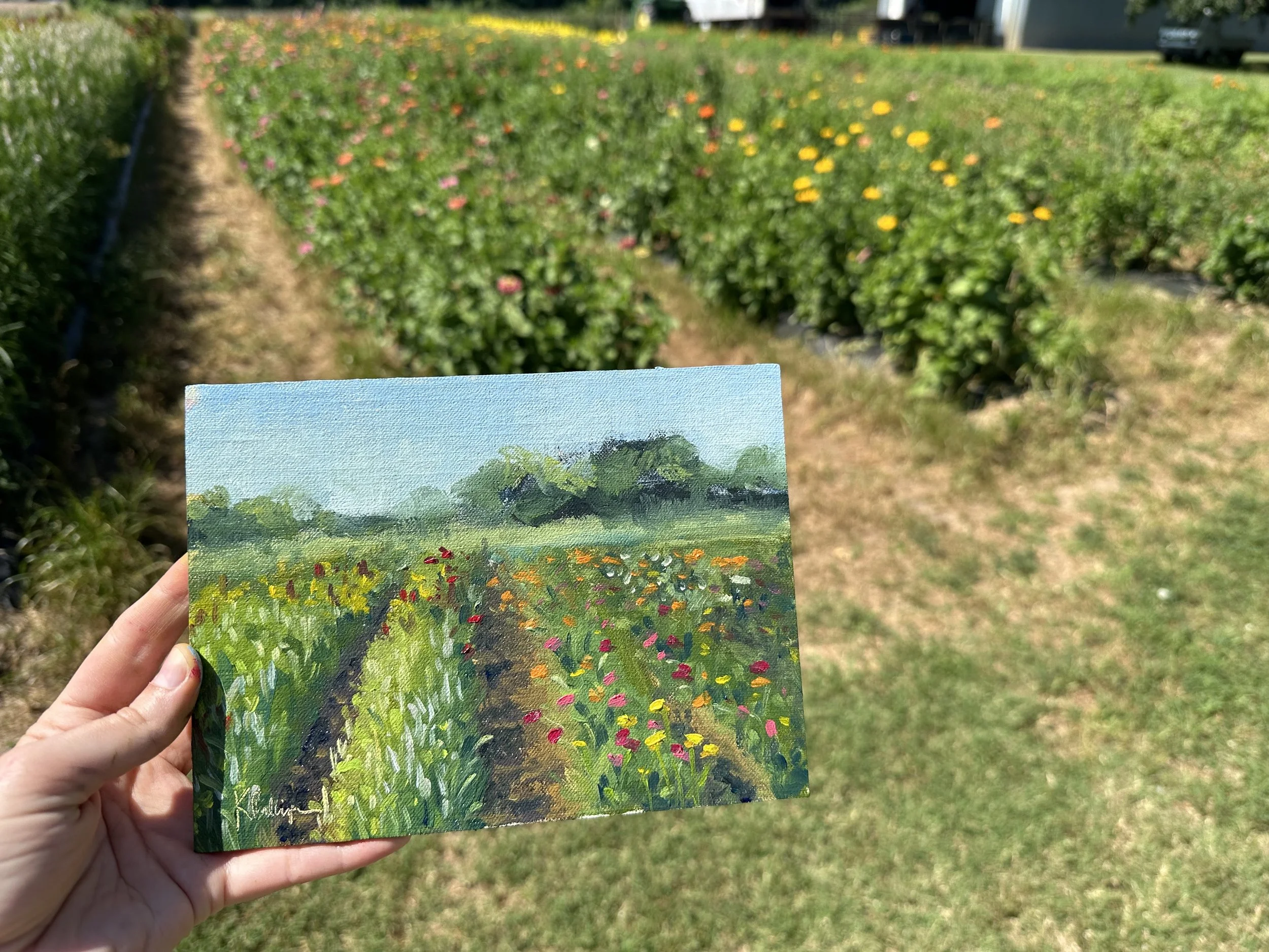 A person holding a small painted landscape in front of a real garden with rows of colorful flowers and green grass.