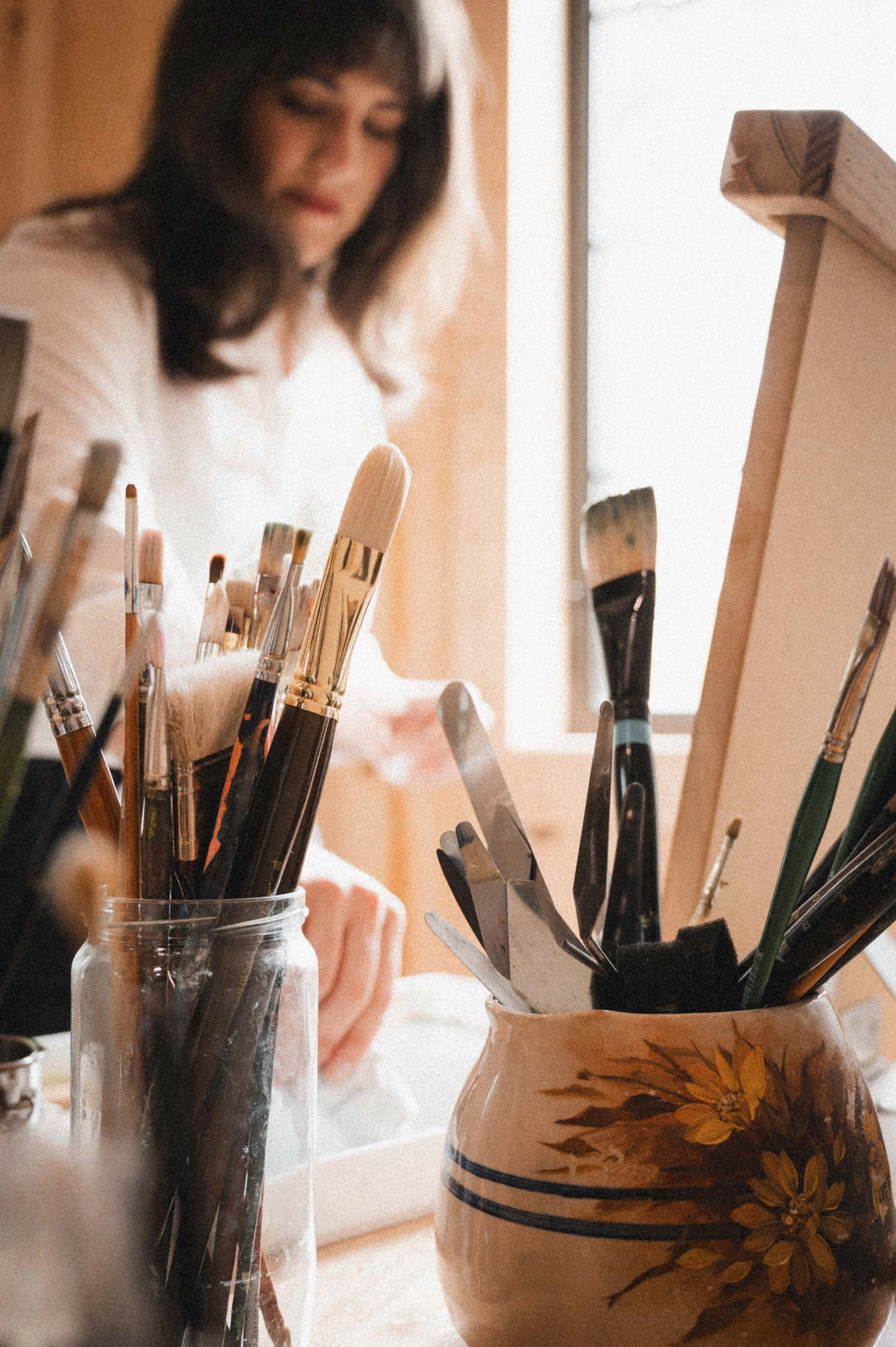A woman is painting, with a focus on her workspace which includes jars filled with paintbrushes and painting tools. She is near a window, and her face is slightly out of focus in the background.