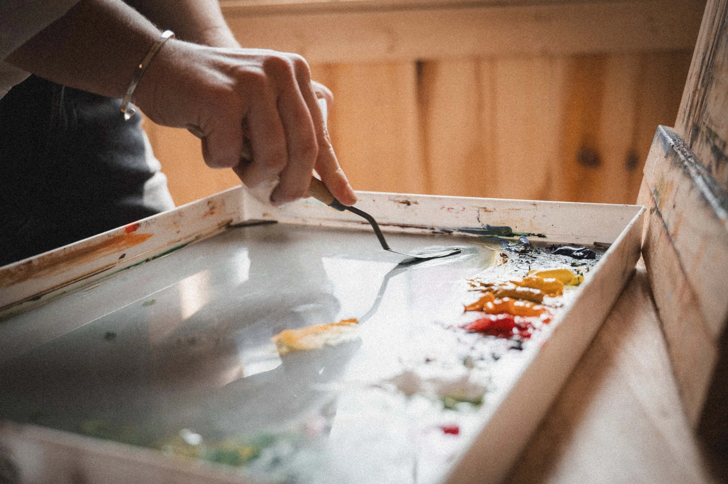 Close-up of a person's hand mixing paints with a palette knife on a painter's palette, with various colors of paint on the palette and a wooden background.