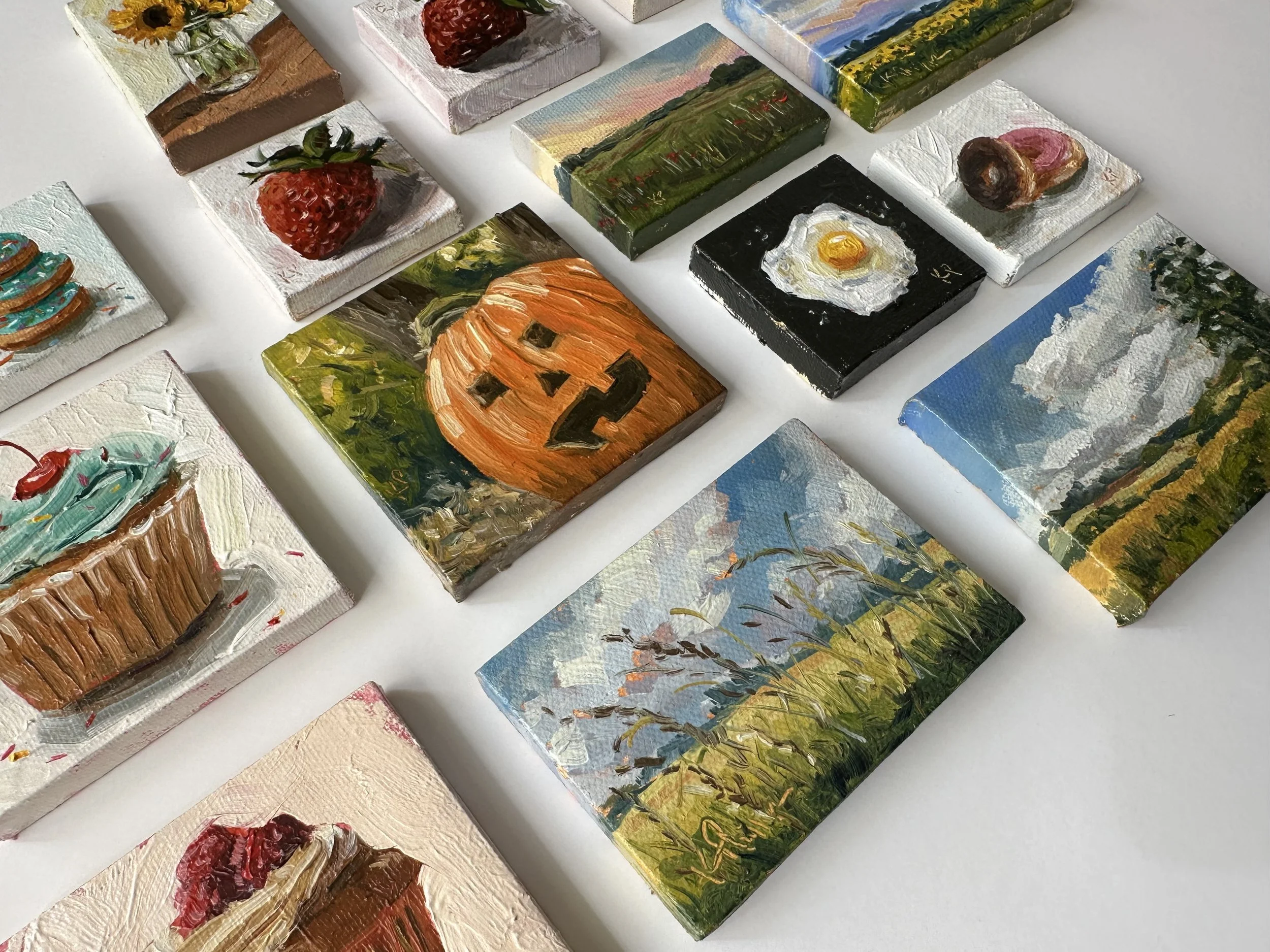 Collection of small, painted canvases featuring various themes like landscape, food, and holiday images, displayed on a white surface.