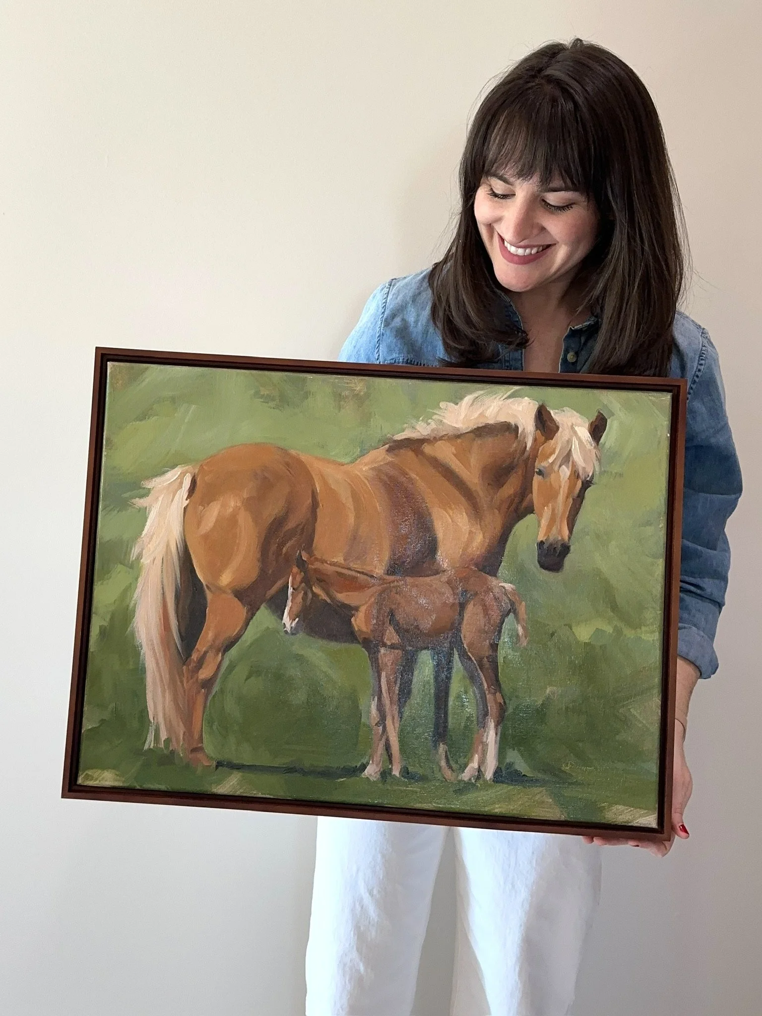 A woman with dark hair and a denim shirt holding a framed painting of a mare and a foal standing on green grass.