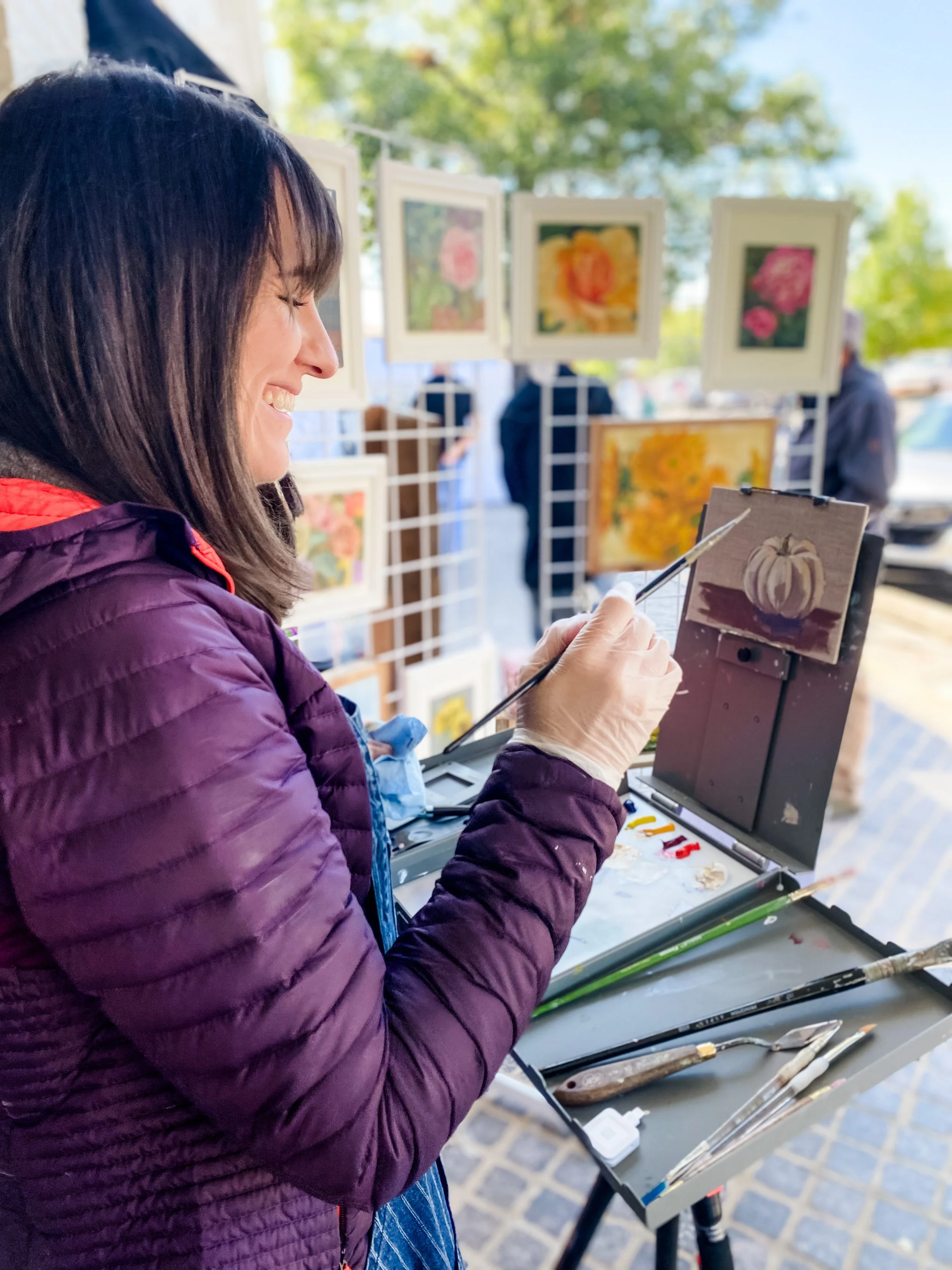 A woman painting a white pumpkin on a small canvas at an outdoor art market, with framed floral paintings displayed on stands in the background.