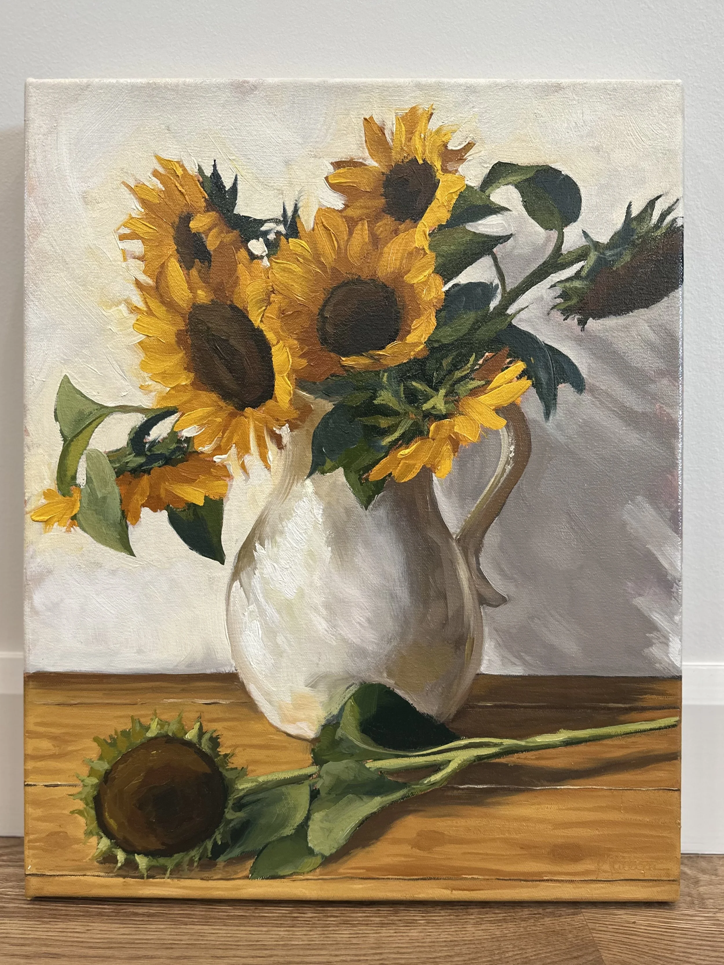Oil painting of a bouquet of sunflowers in a white pitcher, with a single sunflower lying on a wooden table.