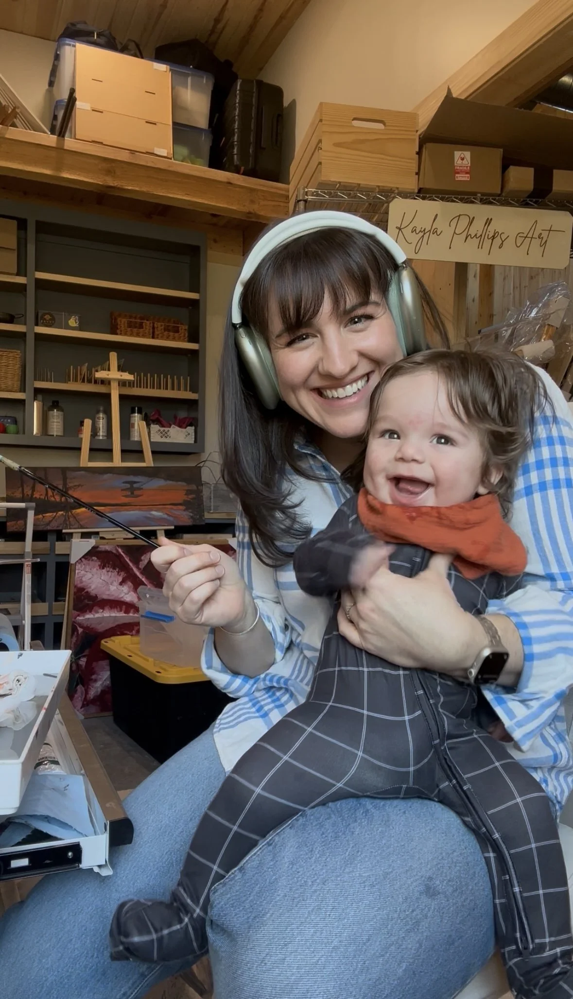 A woman wearing headphones, smiling and holding a young child who is also smiling happily and wearing a colorful outfit with plaid pants, in a craft room with shelves and art supplies.