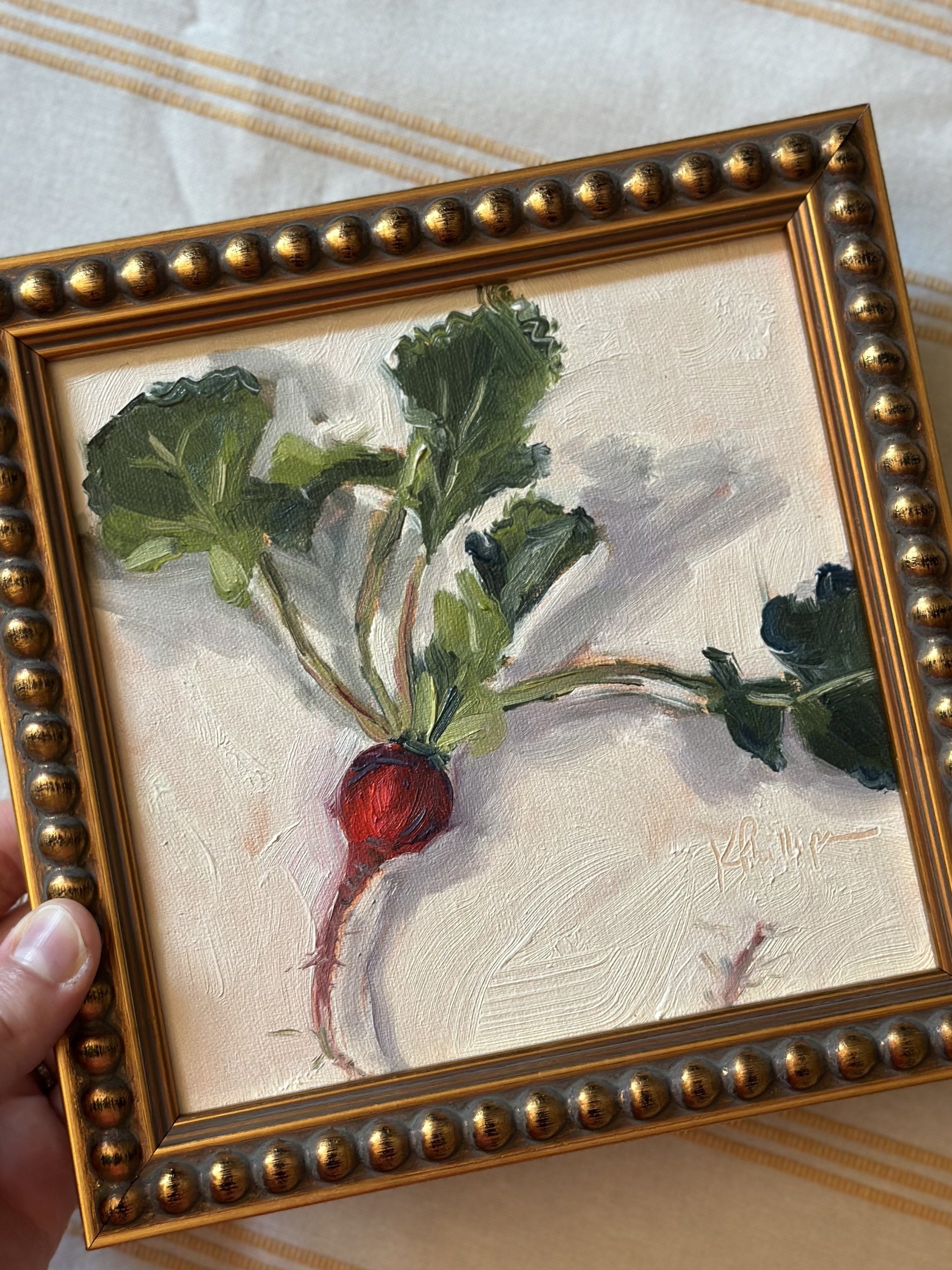 Painted illustration of a radish with green leaves in a gold framed picture, held by a person's hand.