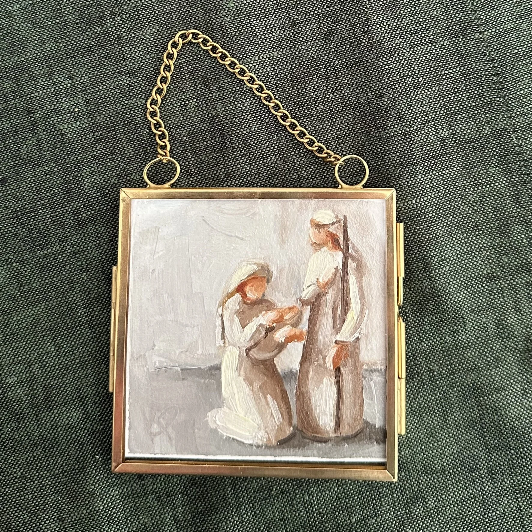 Miniature painting of two women, one kneeling and the other standing, holding something together, with the standing woman wearing a hat, inside a gold frame with a chain, on a dark fabric surface.