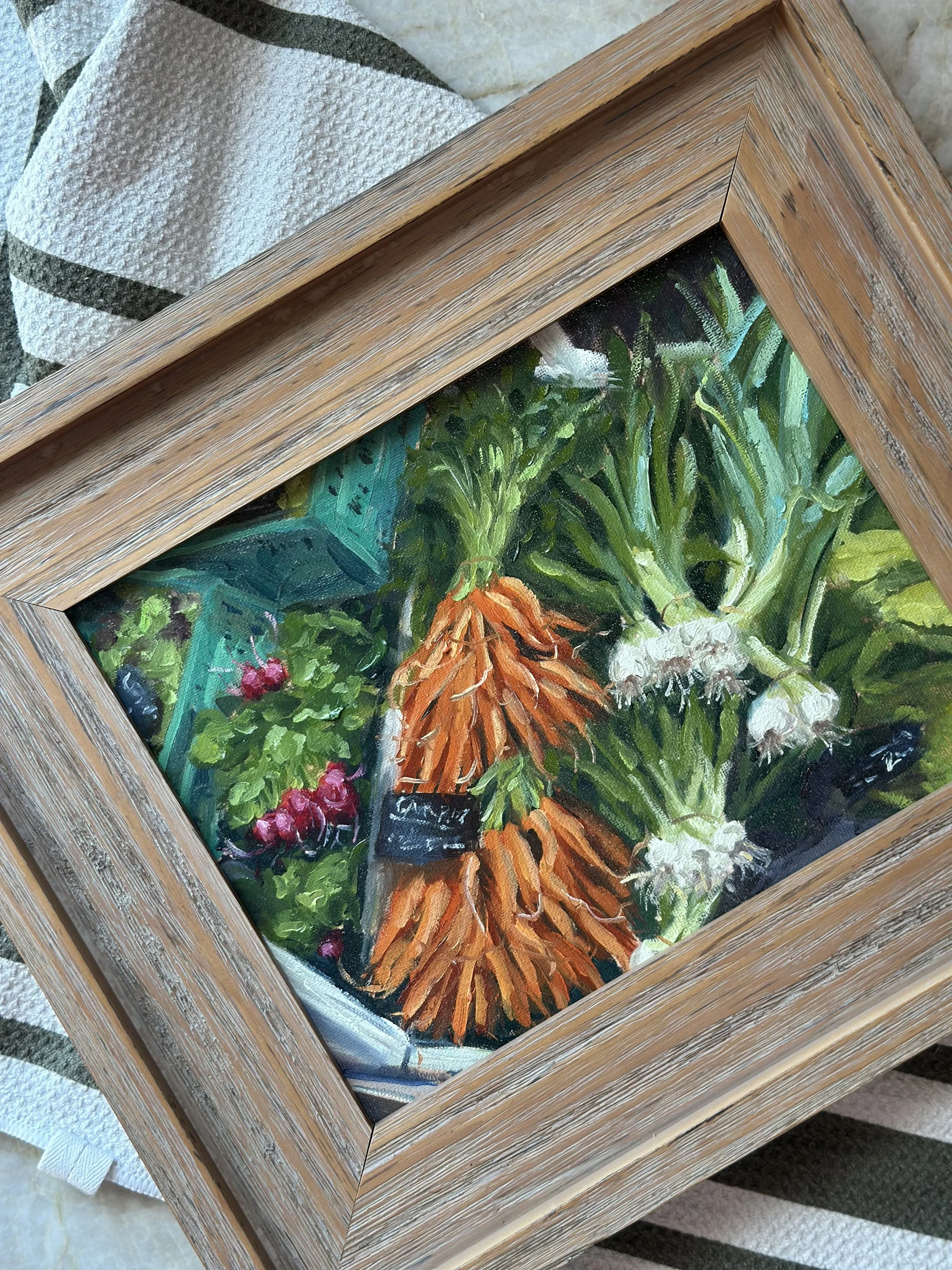 A framed painting of fresh vegetables including carrots, onions, and greens, placed on a cloth surface.