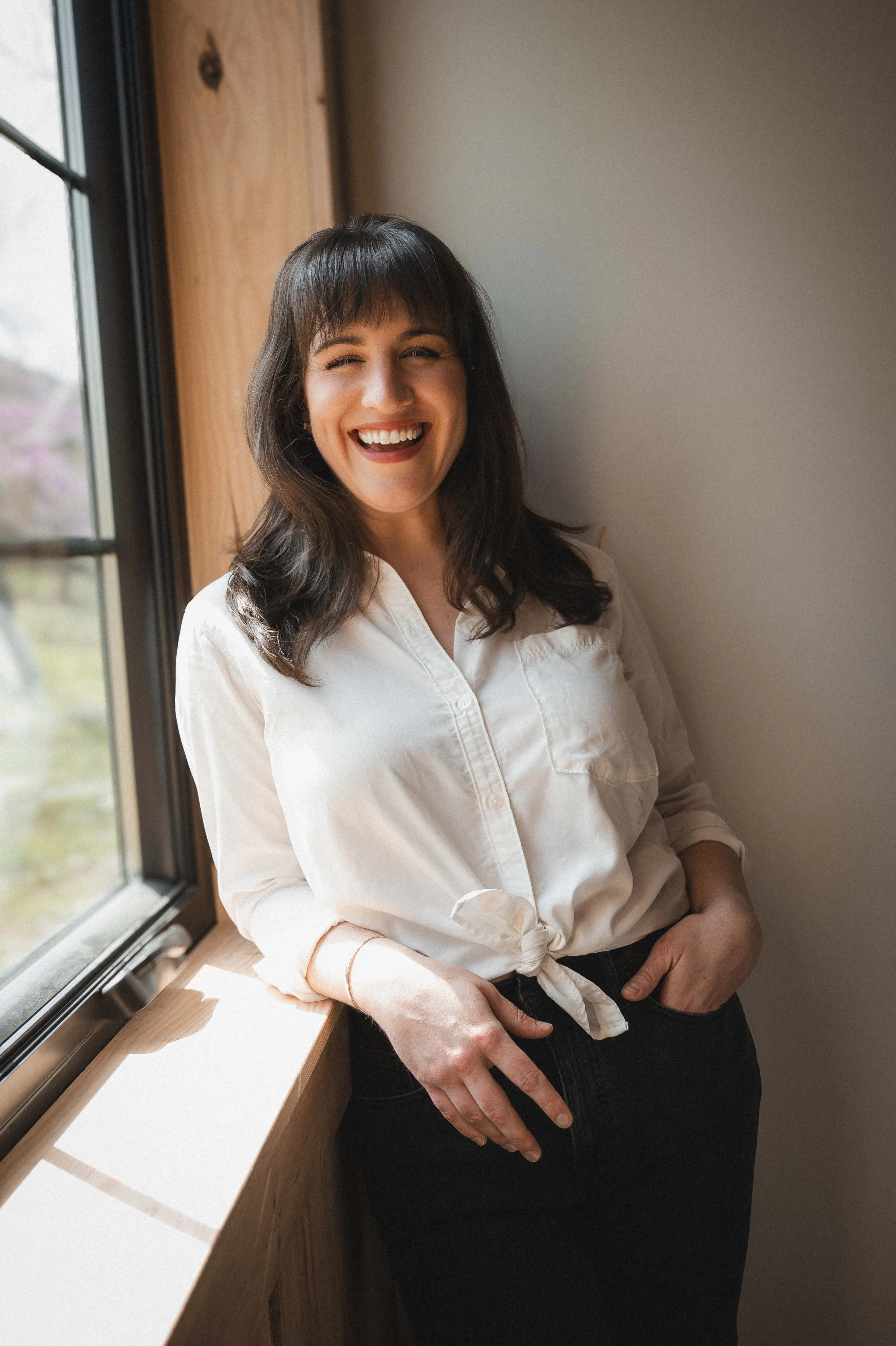 A woman with dark brown hair, wearing a white button-up shirt tied at the waist and black pants, standing next to a window and smiling.