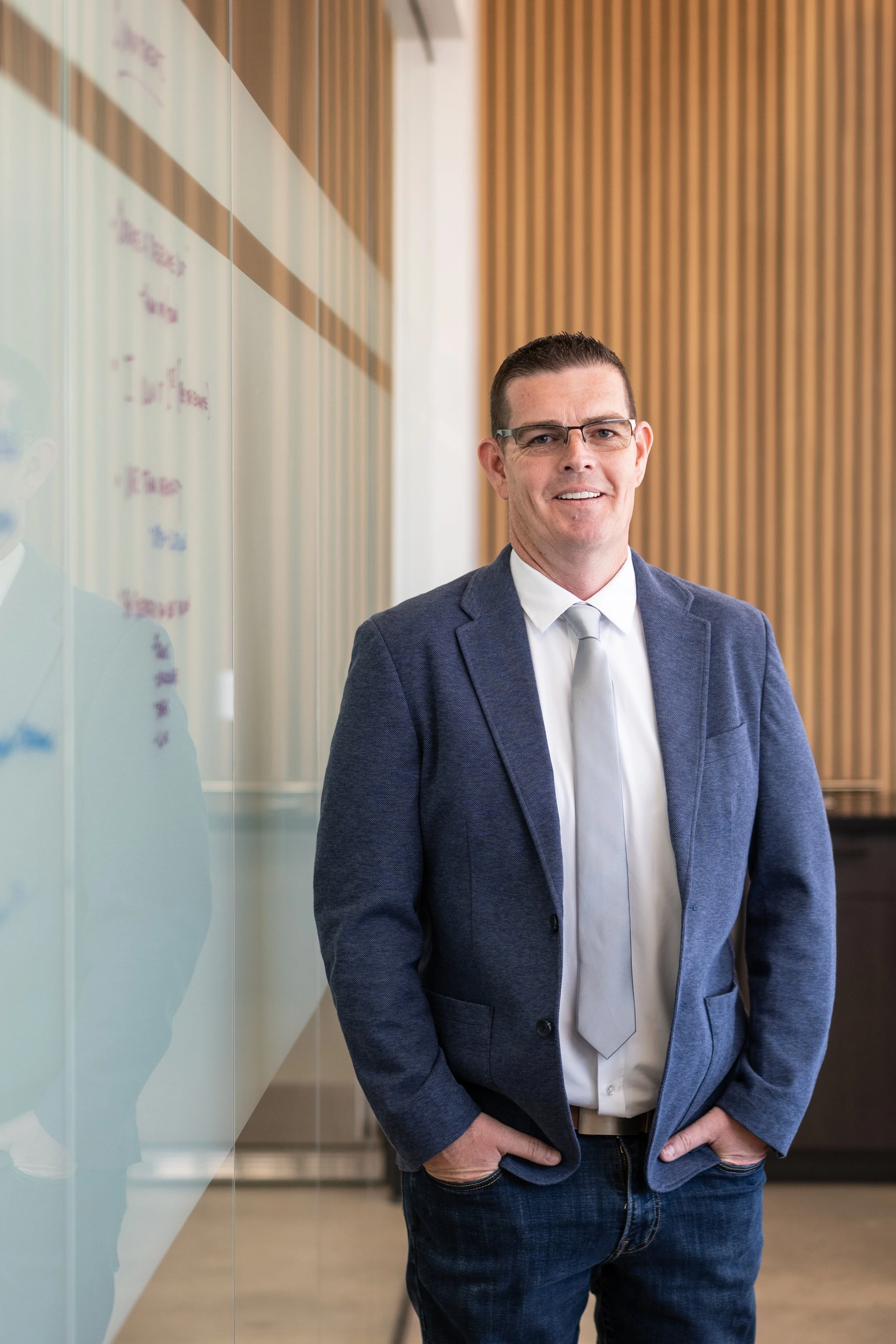 A man in a blue blazer, white shirt, and light-colored tie posing in an office environment with wood paneling and a glass wall.