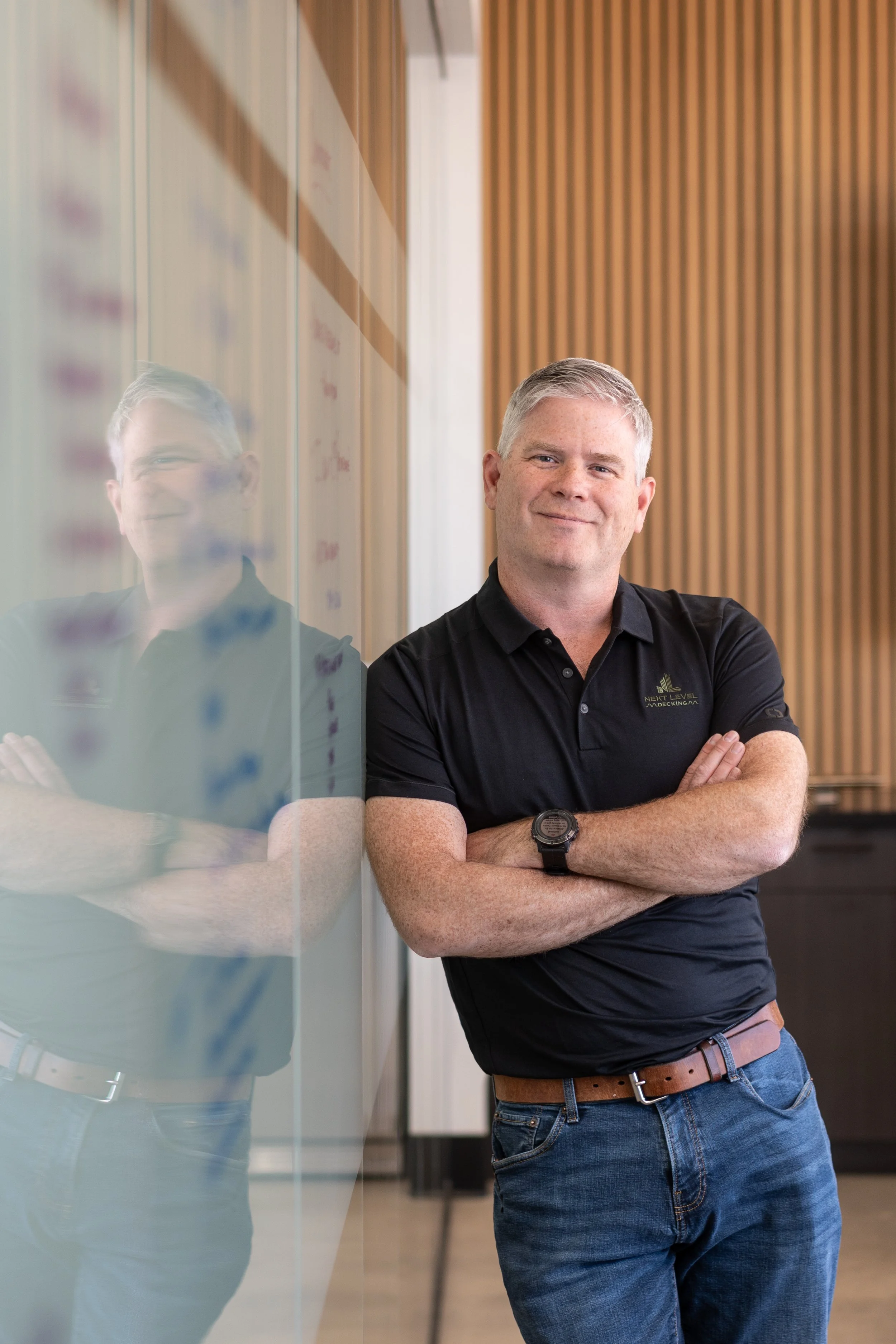 A man leaning against a glass wall, smiling with arms crossed, in a modern office or conference room, wearing a black polo shirt and blue jeans.