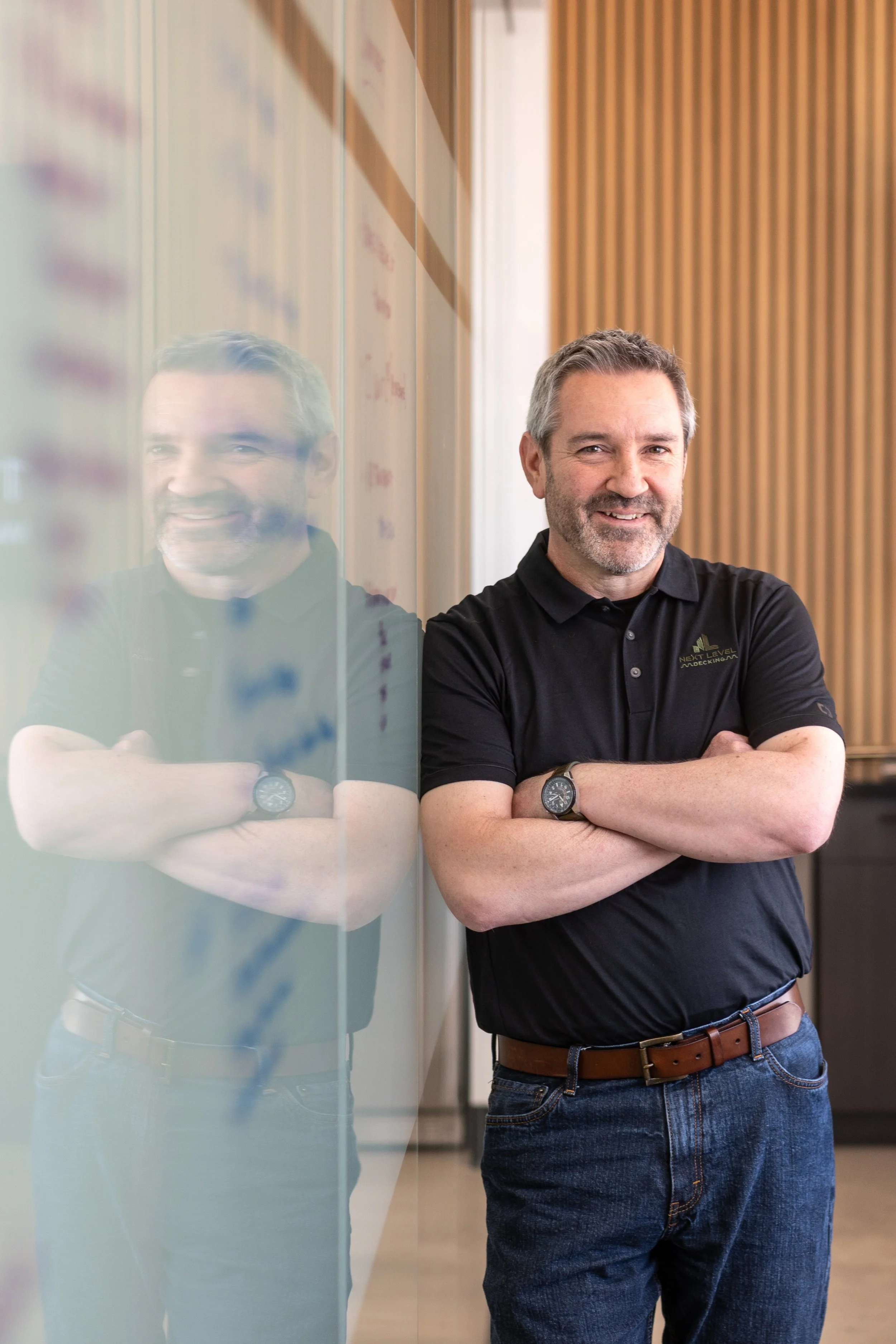 A middle-aged man with gray hair and beard standing with arms crossed, smiling, next to a glass wall showing his reflection, in a modern office space with wood paneling.