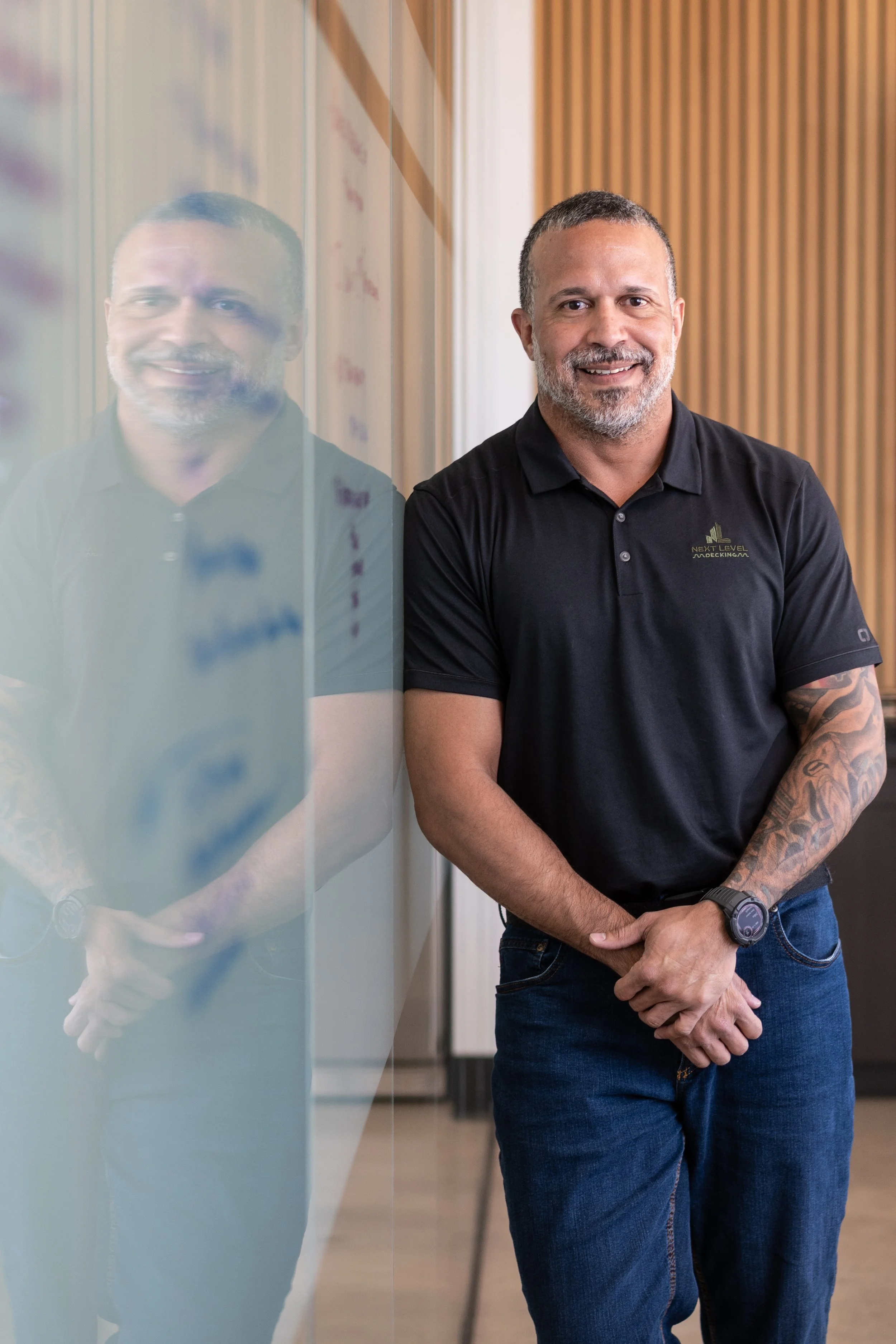 A man in a black polo shirt and jeans standing beside a glass wall, smiling, with his reflection visible on the glass.