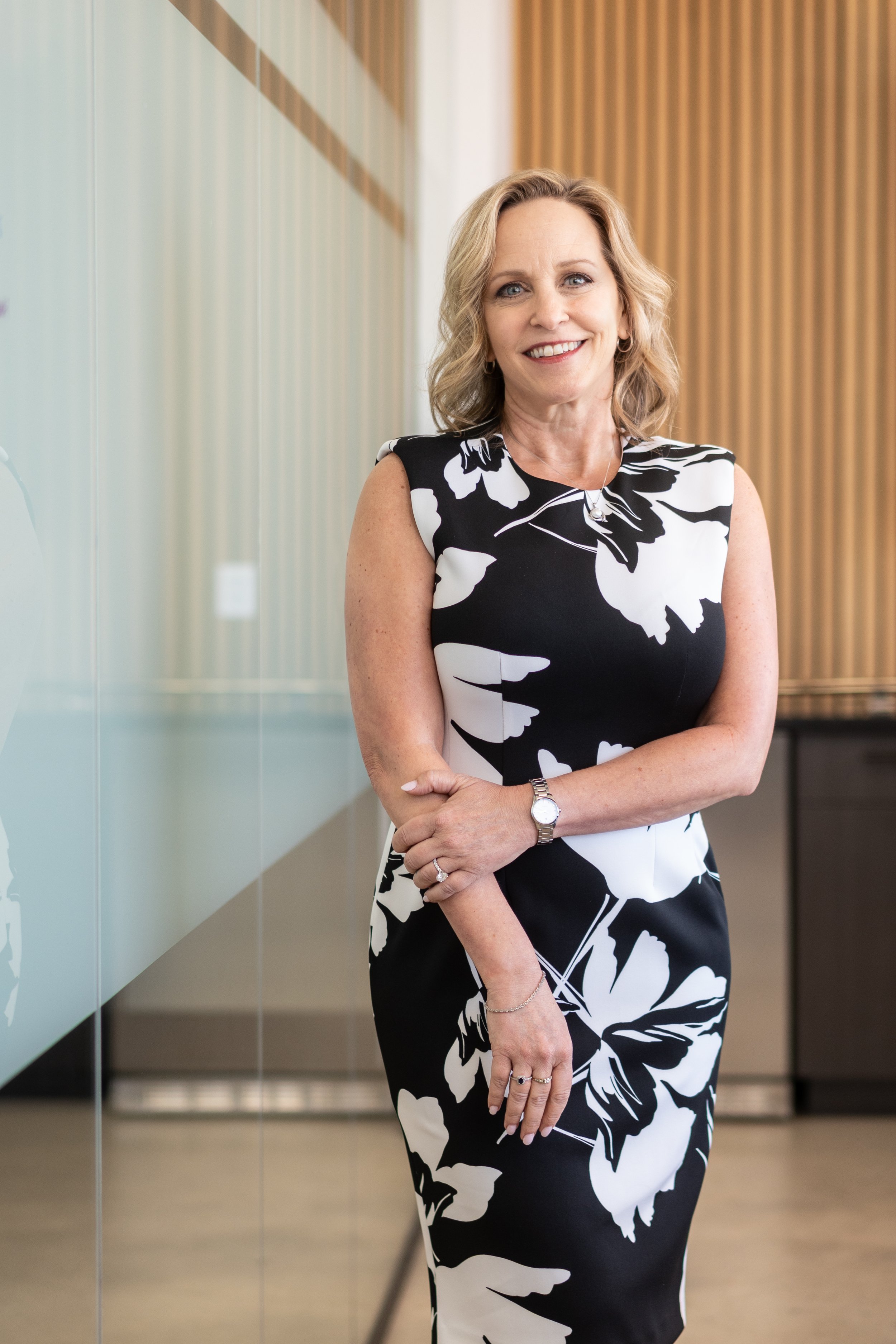 A middle-aged woman with shoulder-length blonde hair in a black and white floral dress standing indoors with in a modern office or lobby setting.