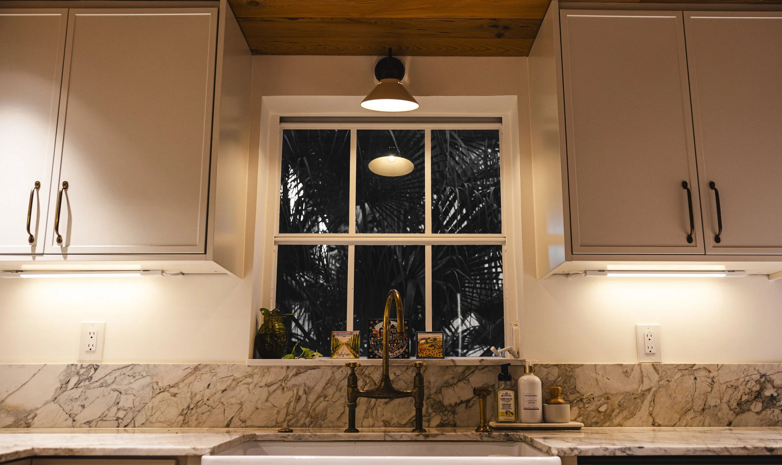 Kitchen sink with brass faucet, marble countertop, window with black and white tropical leaves, overhead light, white cabinets, and various small decor items.