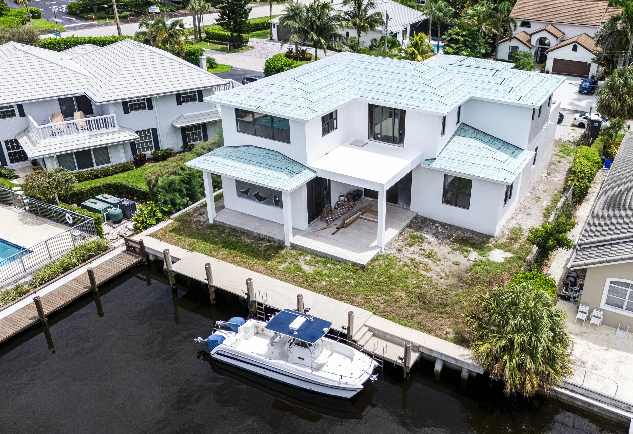 A house under construction beside a canal with a docked boat, surrounded by other residential buildings and greenery.