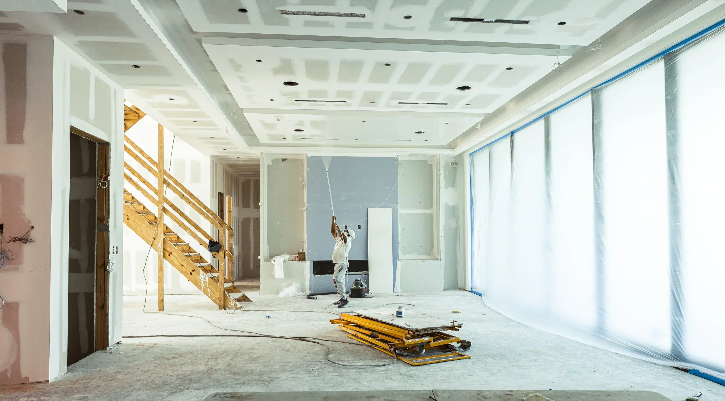 An interior under construction with drywall being installed, a worker painting the wall, wooden stairs on the left, and a large window with plastic sheeting, with construction tools and materials on the floor.