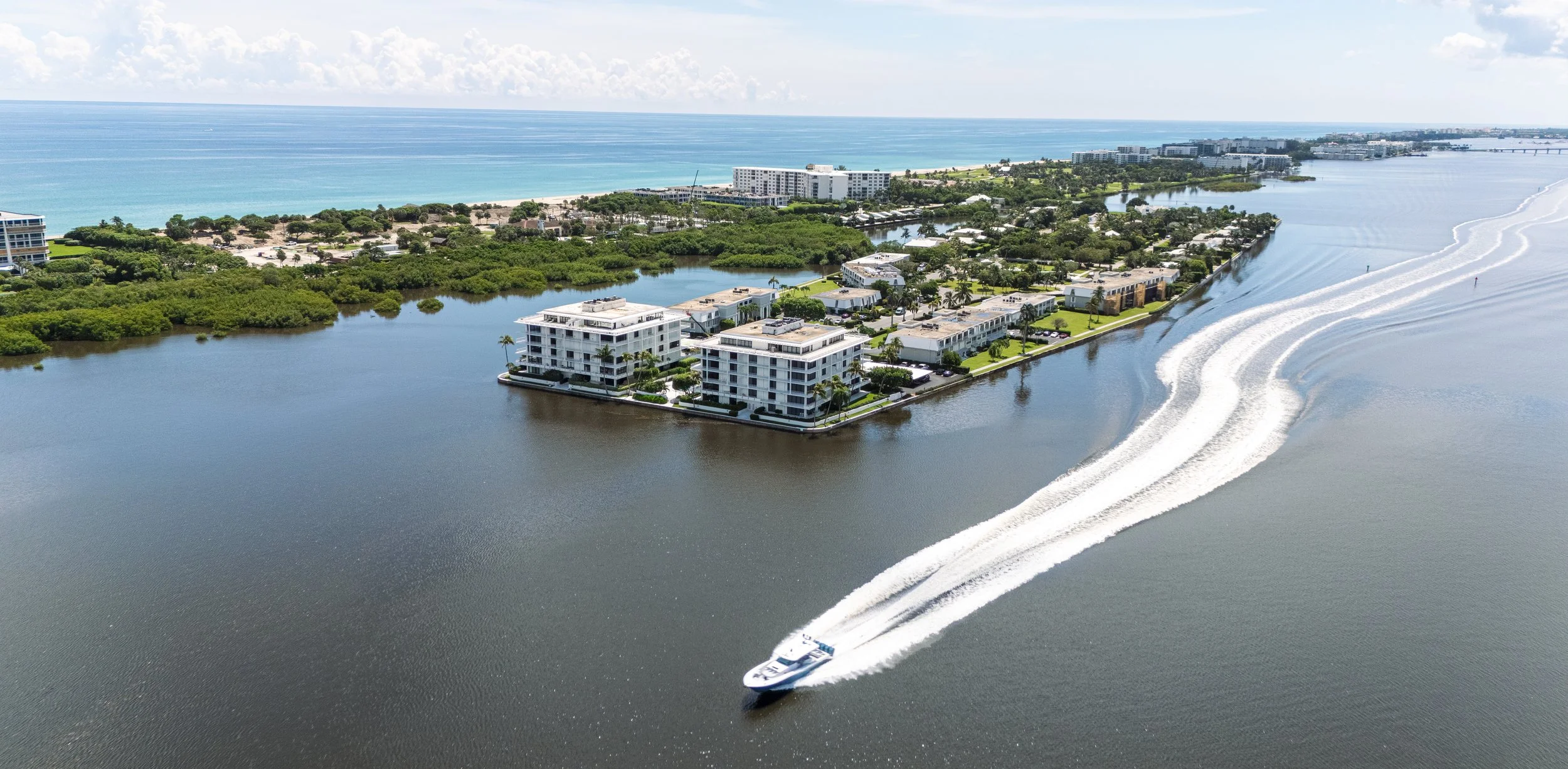 Aerial view of a coastal residential area with modern apartment buildings along the water's edge, a boat cruising through the water creating wakes, and the ocean in the background.