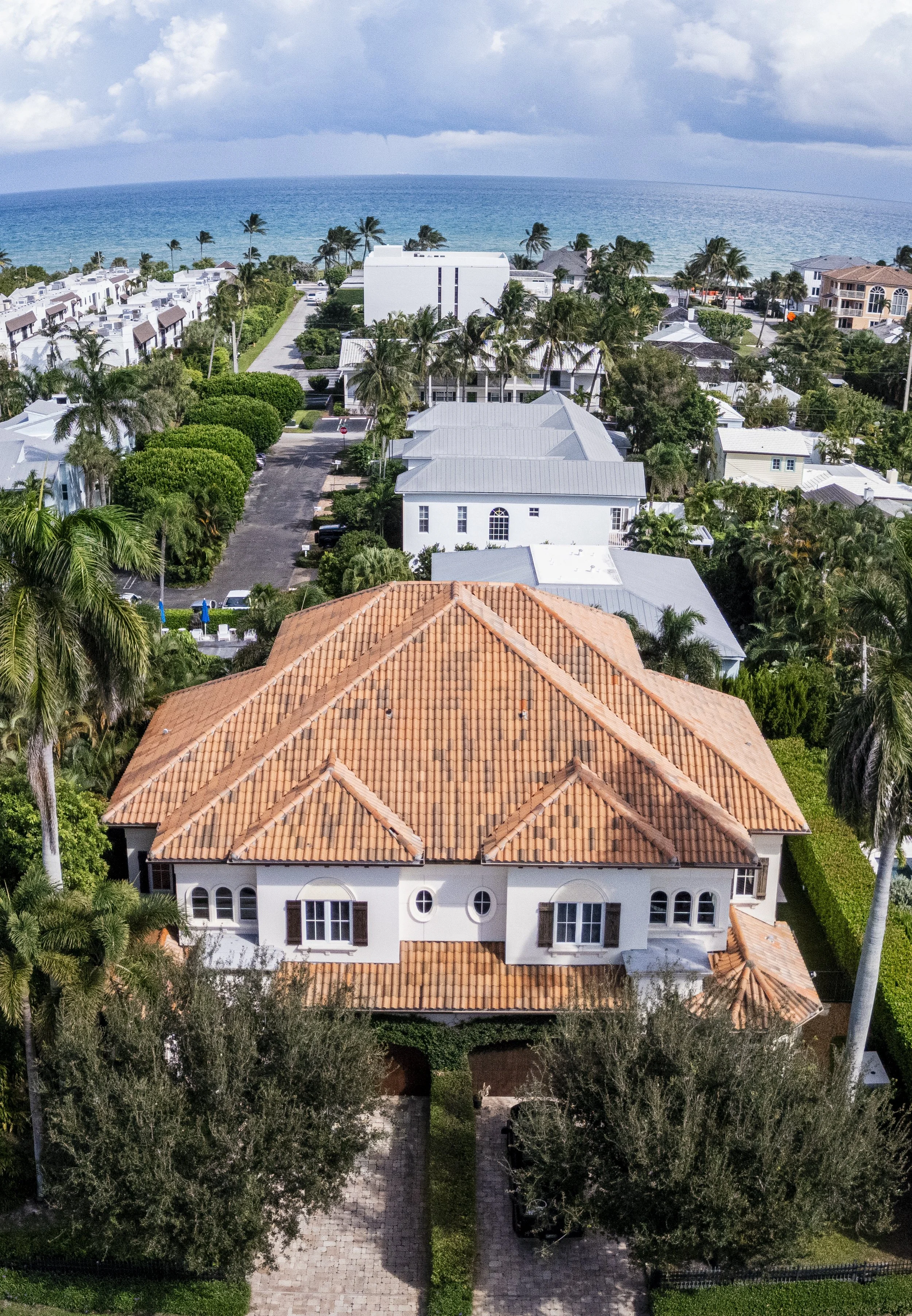 Aerial view of a coastal residential neighborhood with a red-tile roof house in the foreground, surrounded by palm trees, white and gray houses, green vegetation, and the ocean in the background.