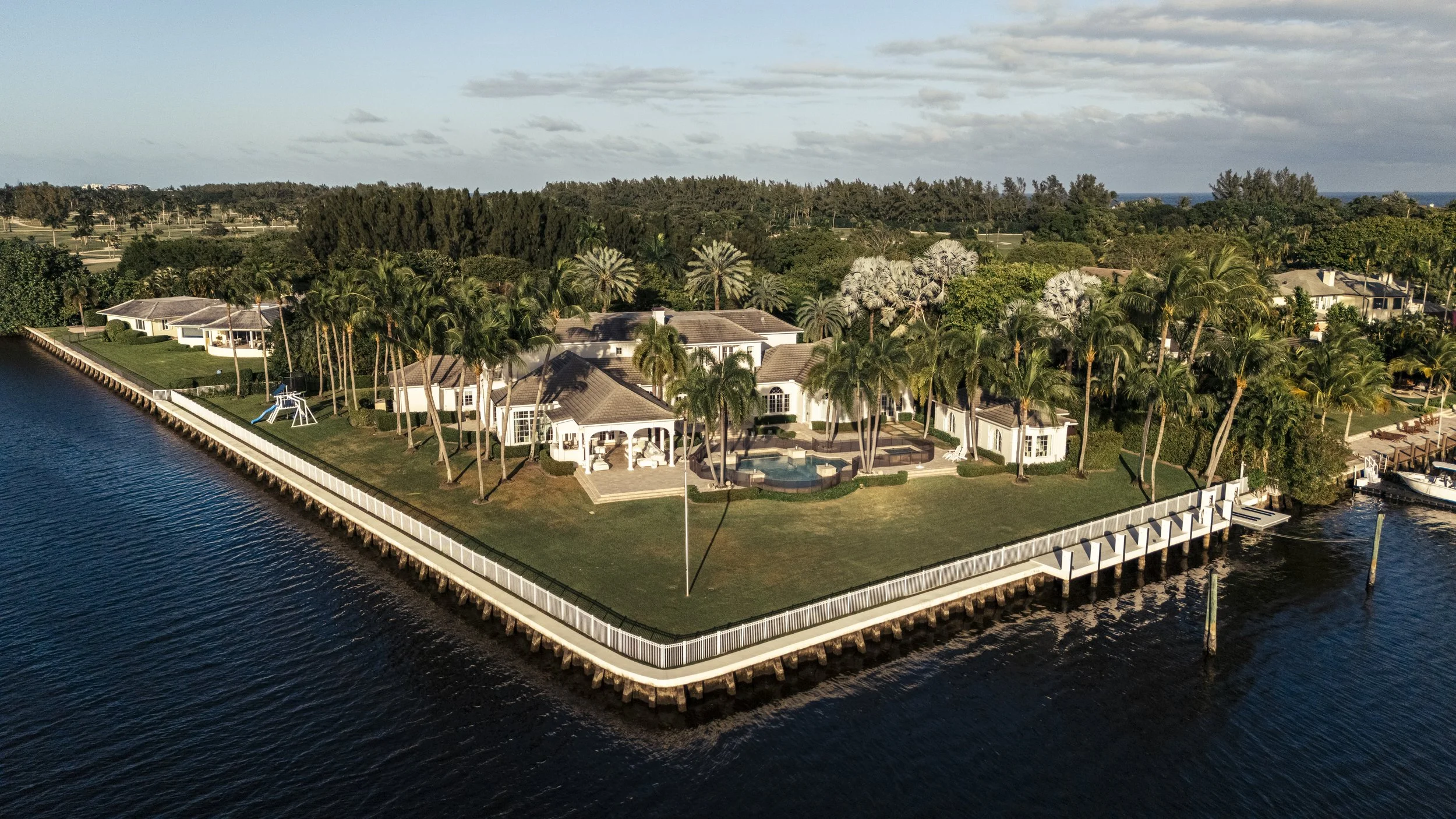 Aerial view of a large waterfront mansion with a swimming pool and palm trees, situated on a grassy lot surrounded by water.