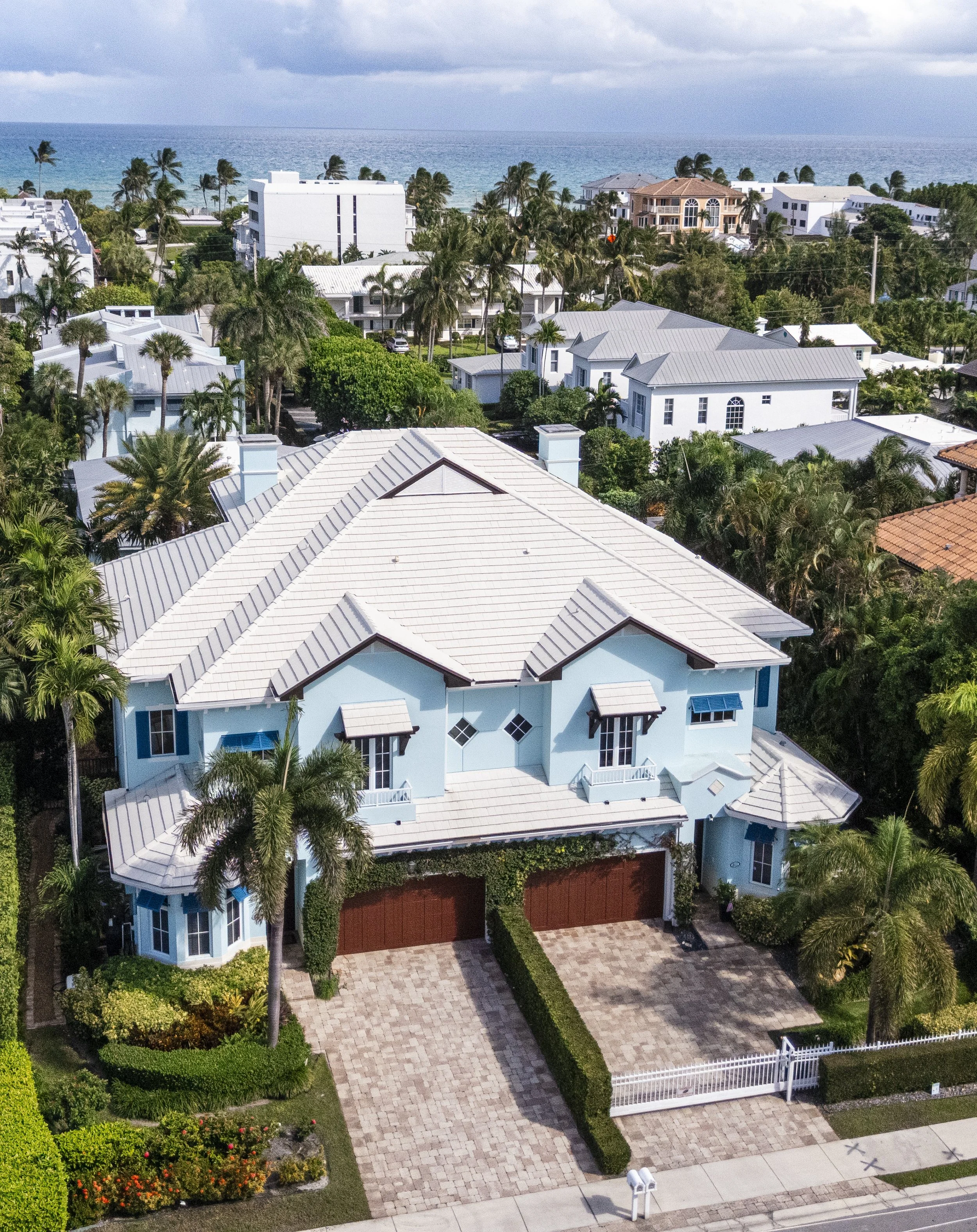 A large, light blue house with a metal roof, surrounded by palm trees, with a driveway leading to a two-car garage, and a view of the ocean in the background.