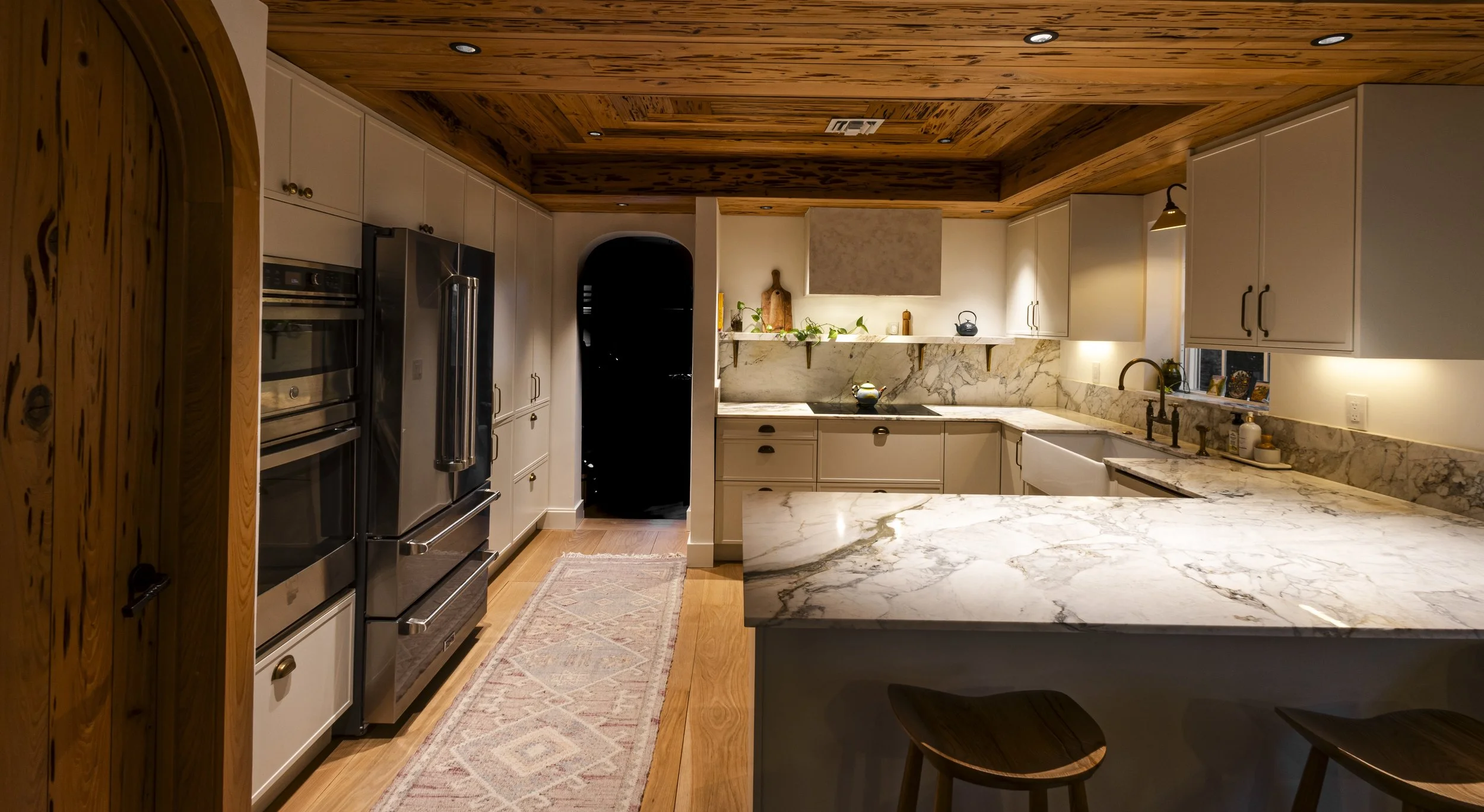 Kitchen with white cabinetry, marble countertops, and a wooden ceiling. Features include a stainless steel fridge, oven, and a marble island with two wooden barstools.