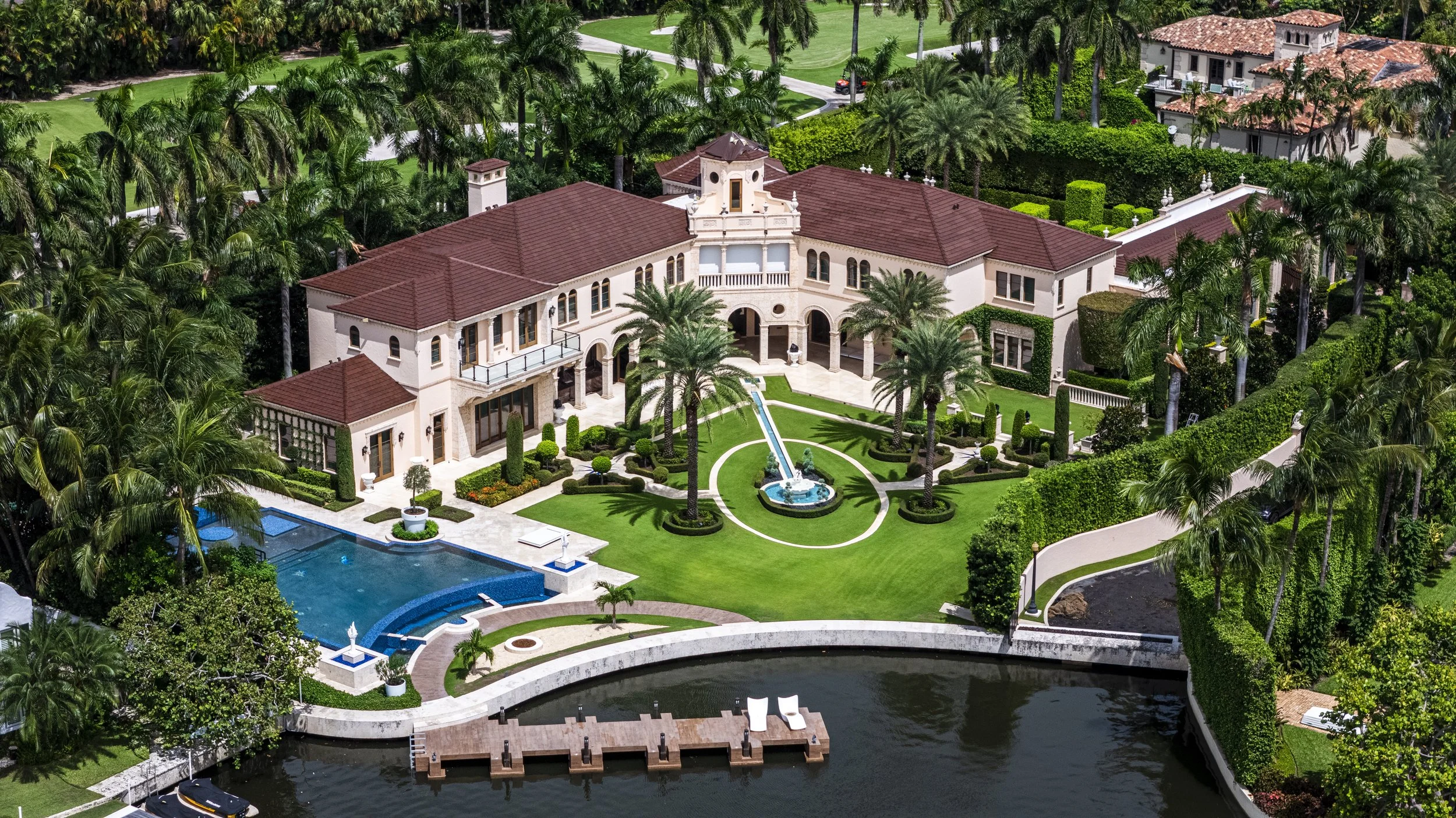 A large mansion with a red-tiled roof, surrounded by lush palm trees and greenery. The property features a swimming pool, a water fountain with a stream, and a dock on a waterway.