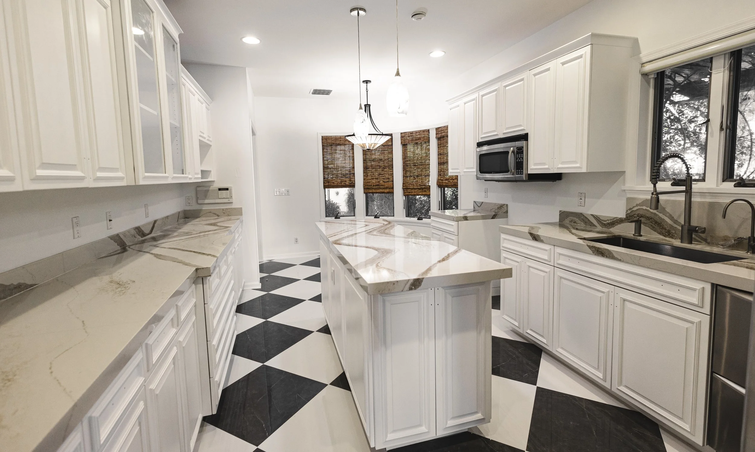 Modern kitchen with white cabinets, marble countertops, black and white checkered floor, windows with woven blinds.