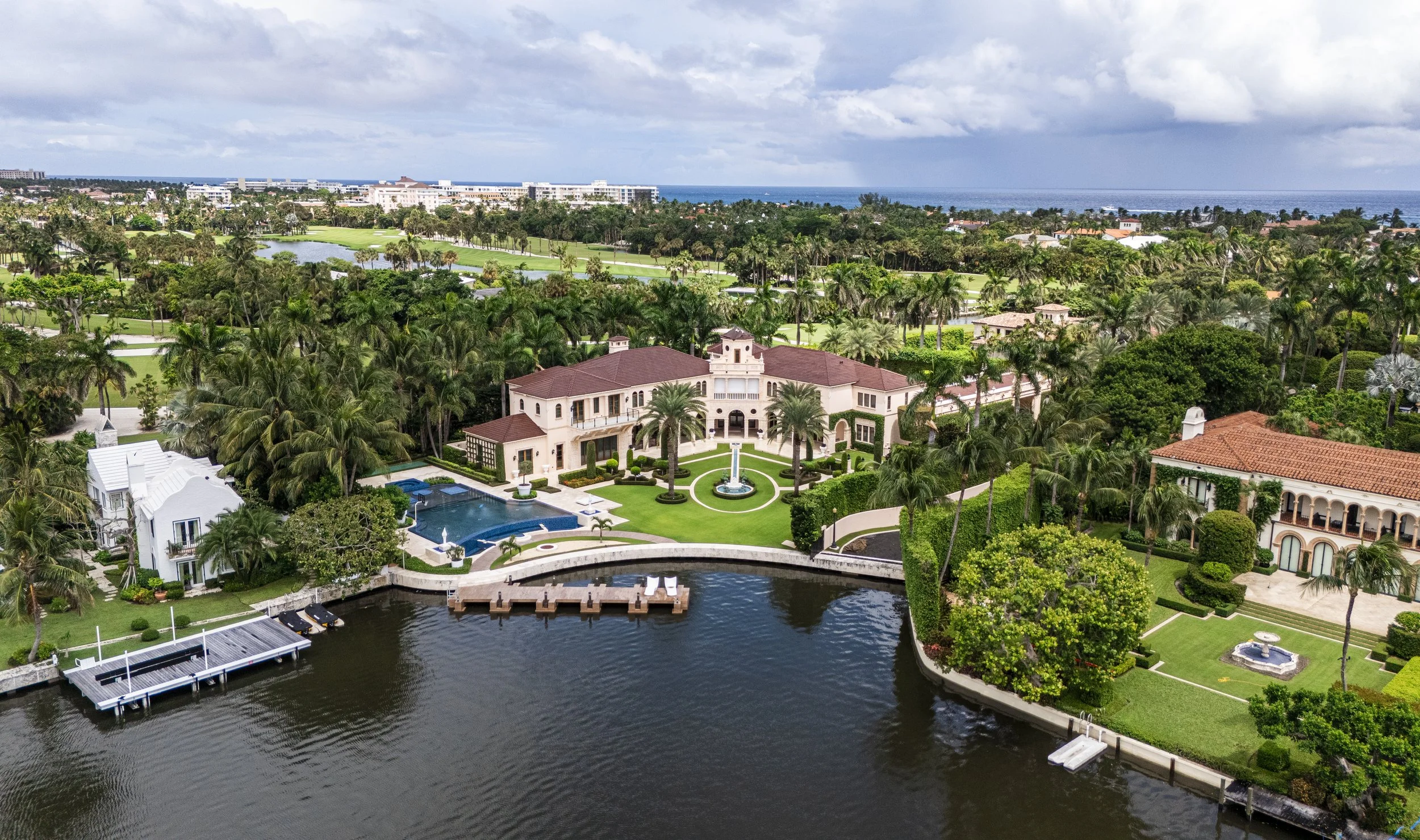 Aerial view of a luxurious waterfront estate with a large mansion, swimming pool, dock, and lush landscaping, overlooking a river with a golf course and distant ocean in the background.