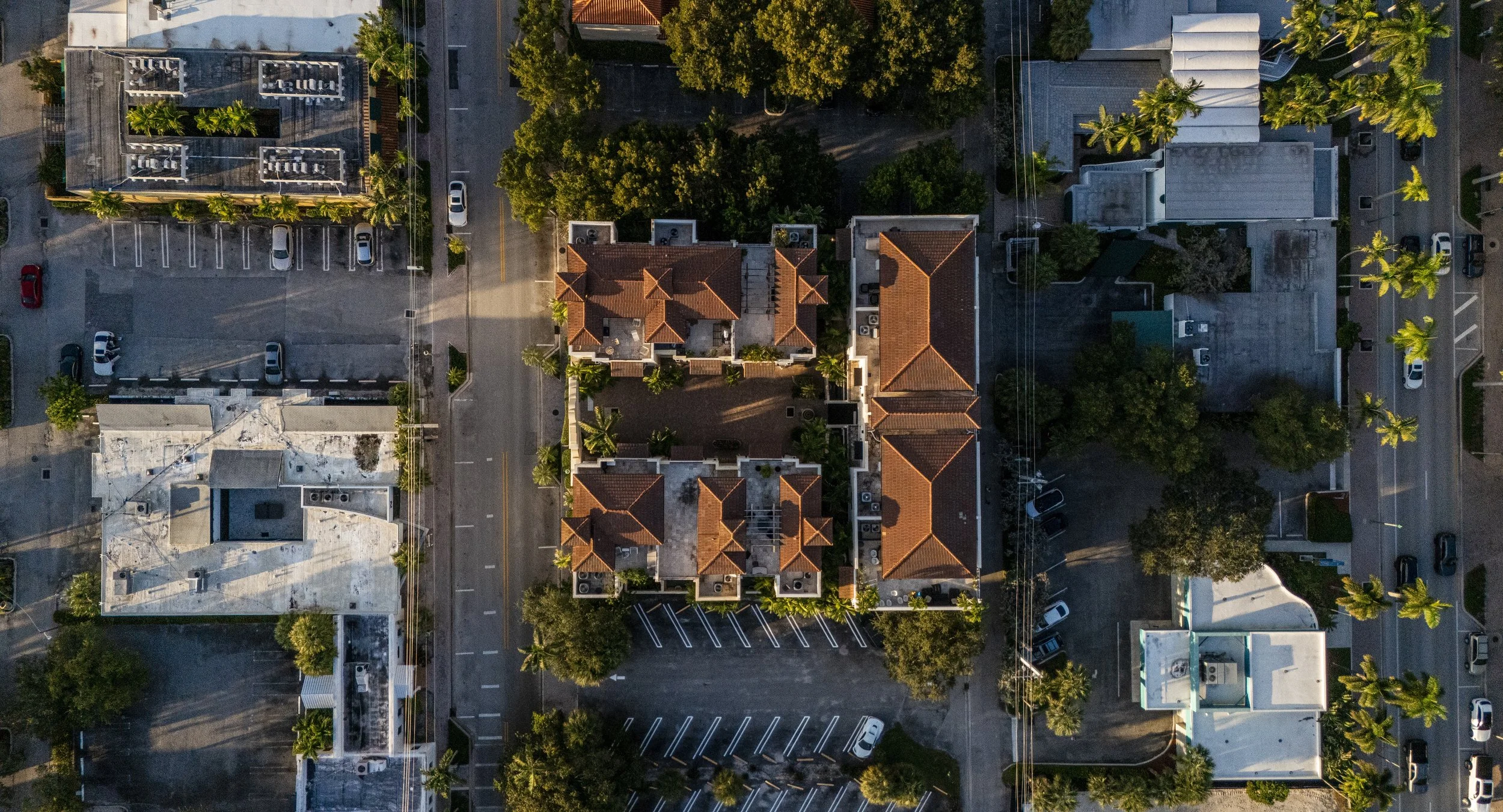 Aerial view of a neighborhood with multiple buildings, parking lots, and trees, taken during the day.