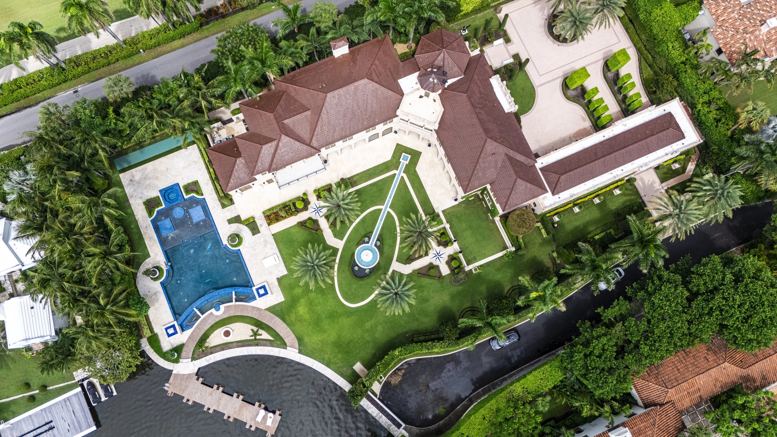 Aerial view of a luxurious mansion with a swimming pool, well-manicured lawn, palm trees, and a long driveway surrounded by greenery.