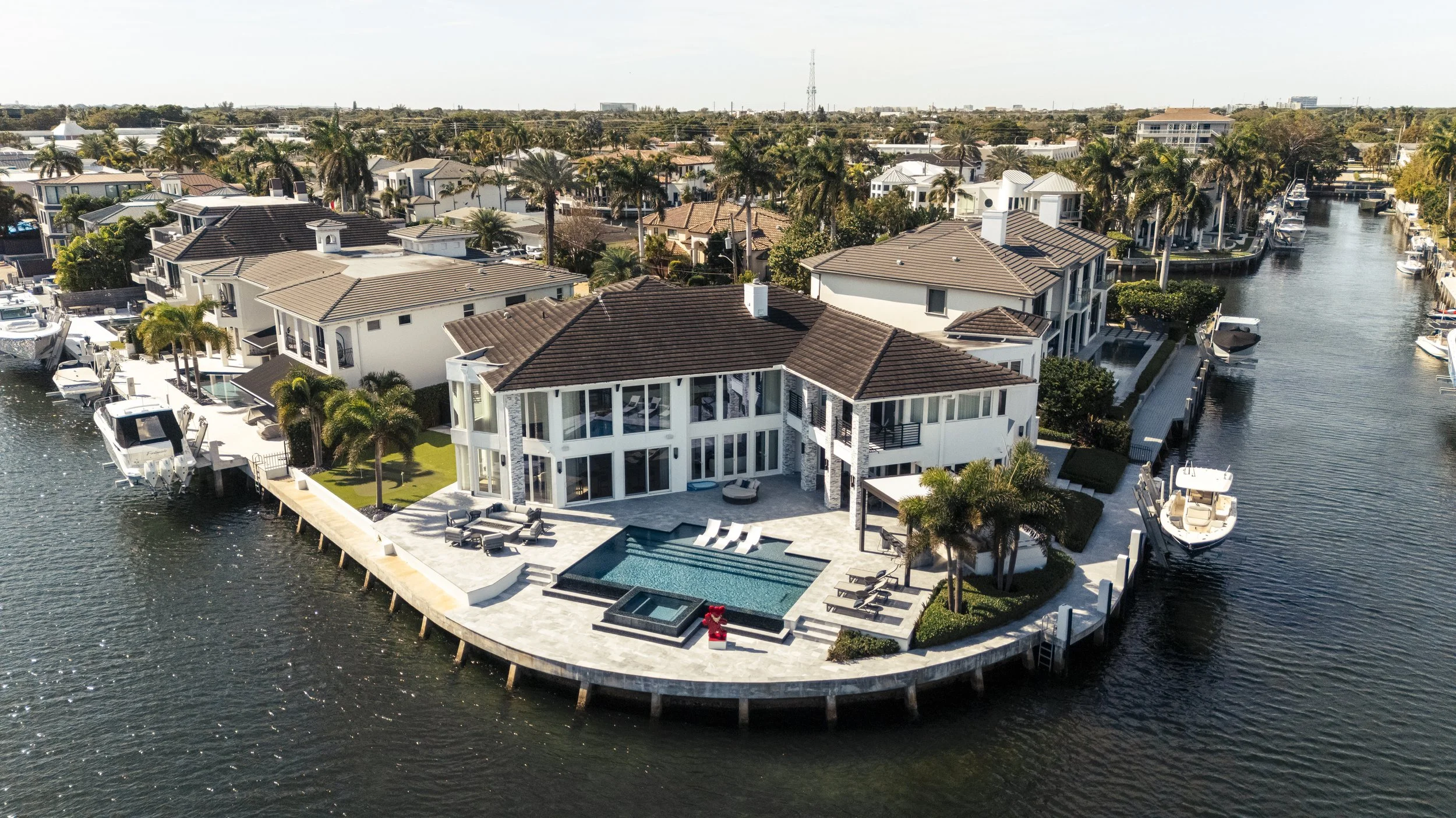 Aerial view of a modern waterfront house with a swimming pool, patio furniture, and several boats docked along the canal in a neighborhood with similar houses.