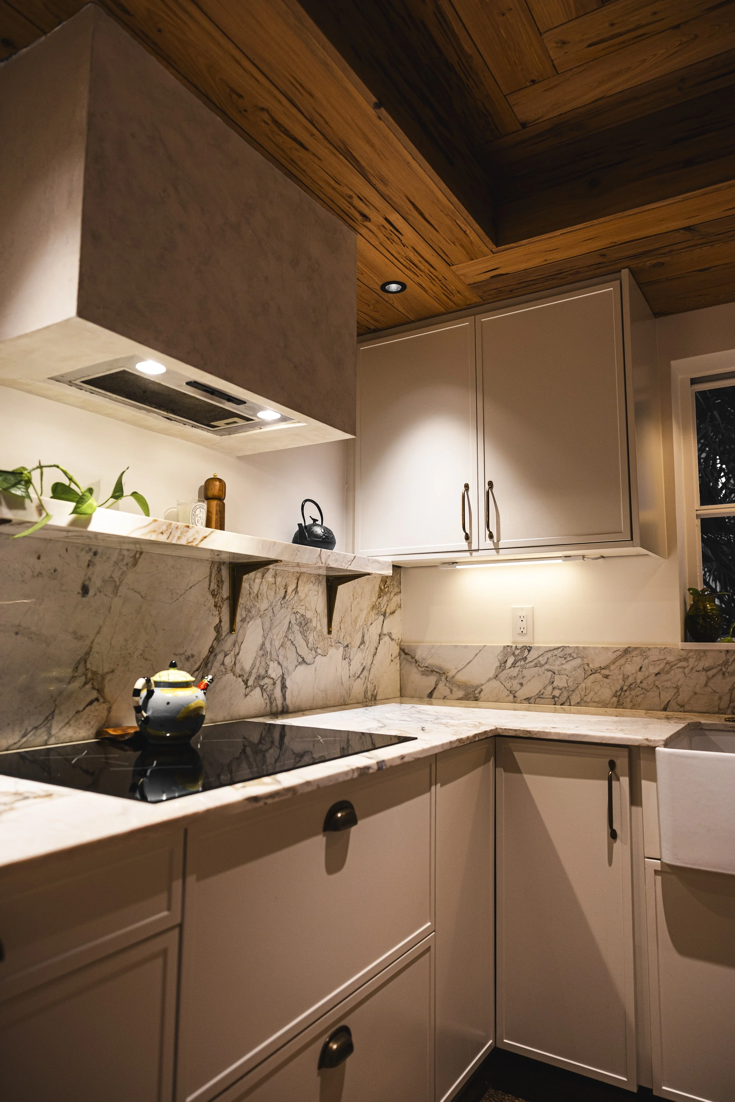 Close-up of a kitchen with marble countertops, white cabinets, wooden ceiling, and a window with night view, with cooking pots, a kettle, and green plants.