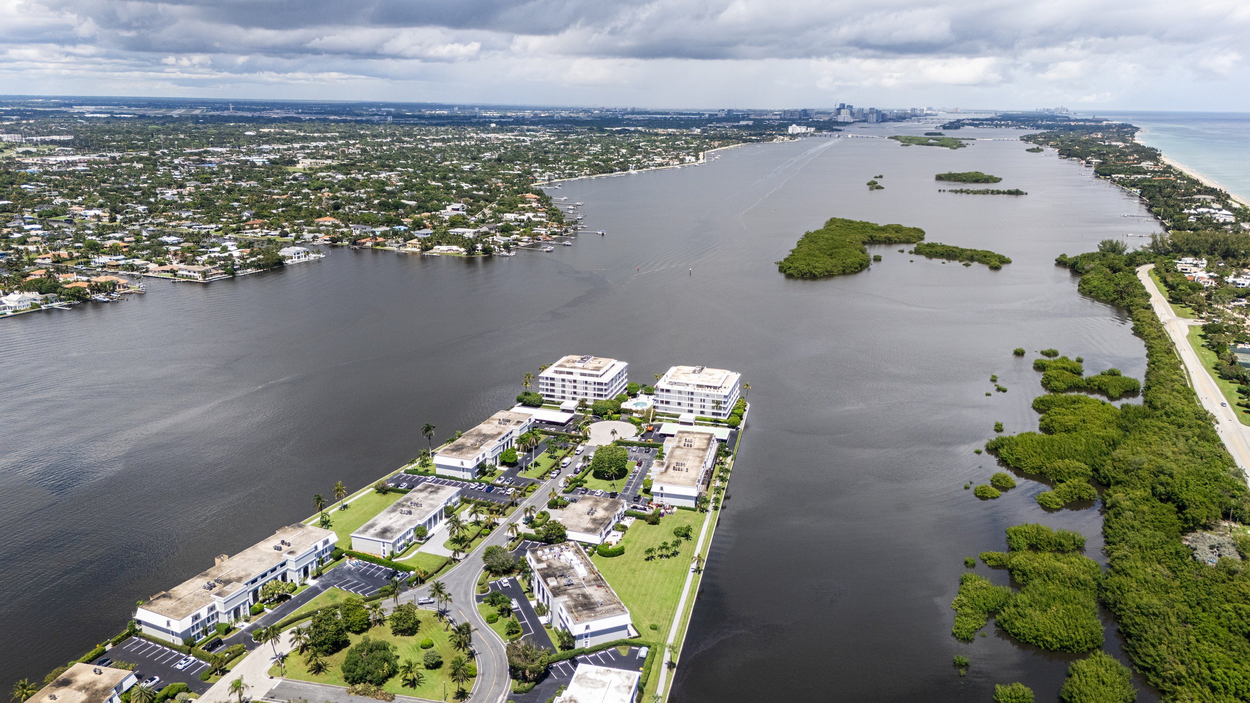 Aerial view of a marina with boats, surrounded by residential buildings, water, and green islands, with a city skyline in the distance under a cloudy sky.