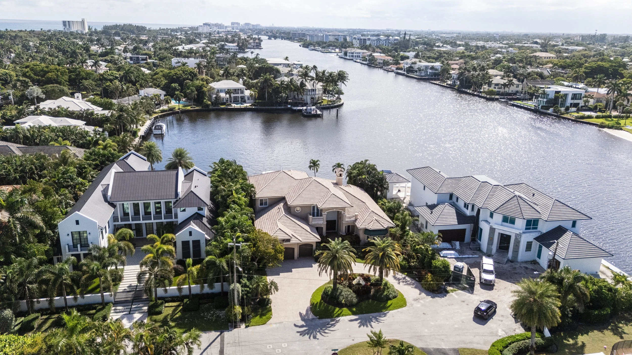 Aerial view of a residential neighborhood with large modern houses along a waterway. The houses are surrounded by lush greenery and palm trees, with boats docked by the water. The waterway extends into the distance, with a cityscape visible on the ho