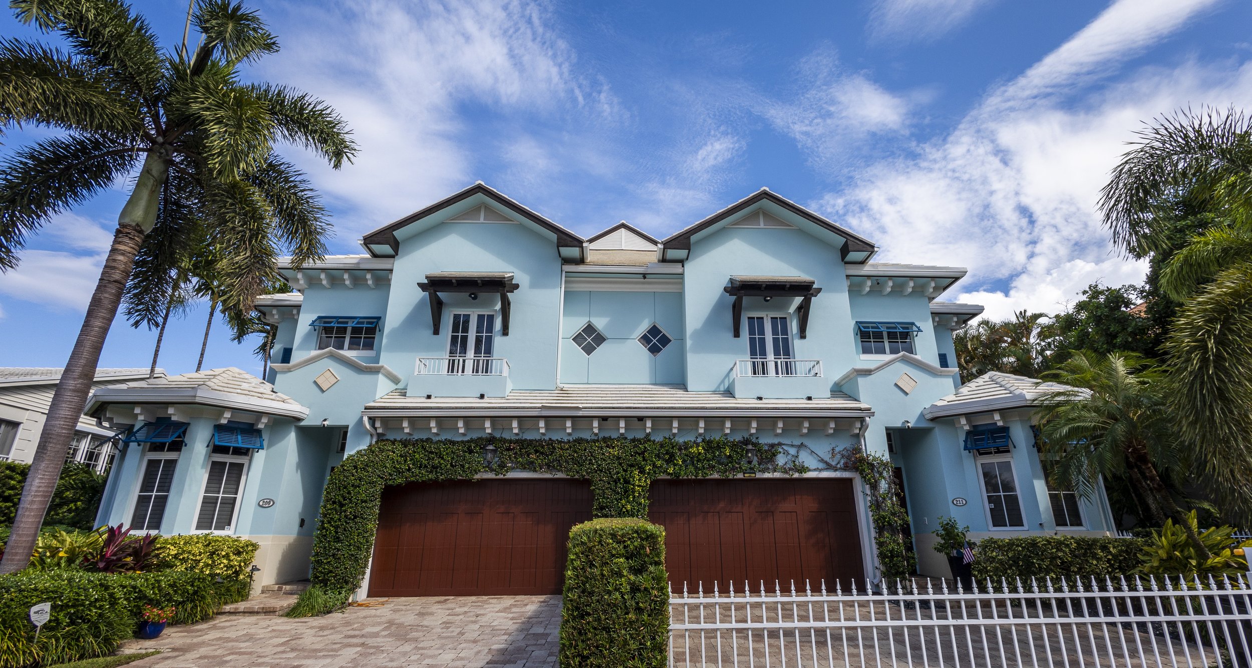 Large blue house with wooden garage doors, surrounded by palm trees and green bushes, under a partly cloudy sky.