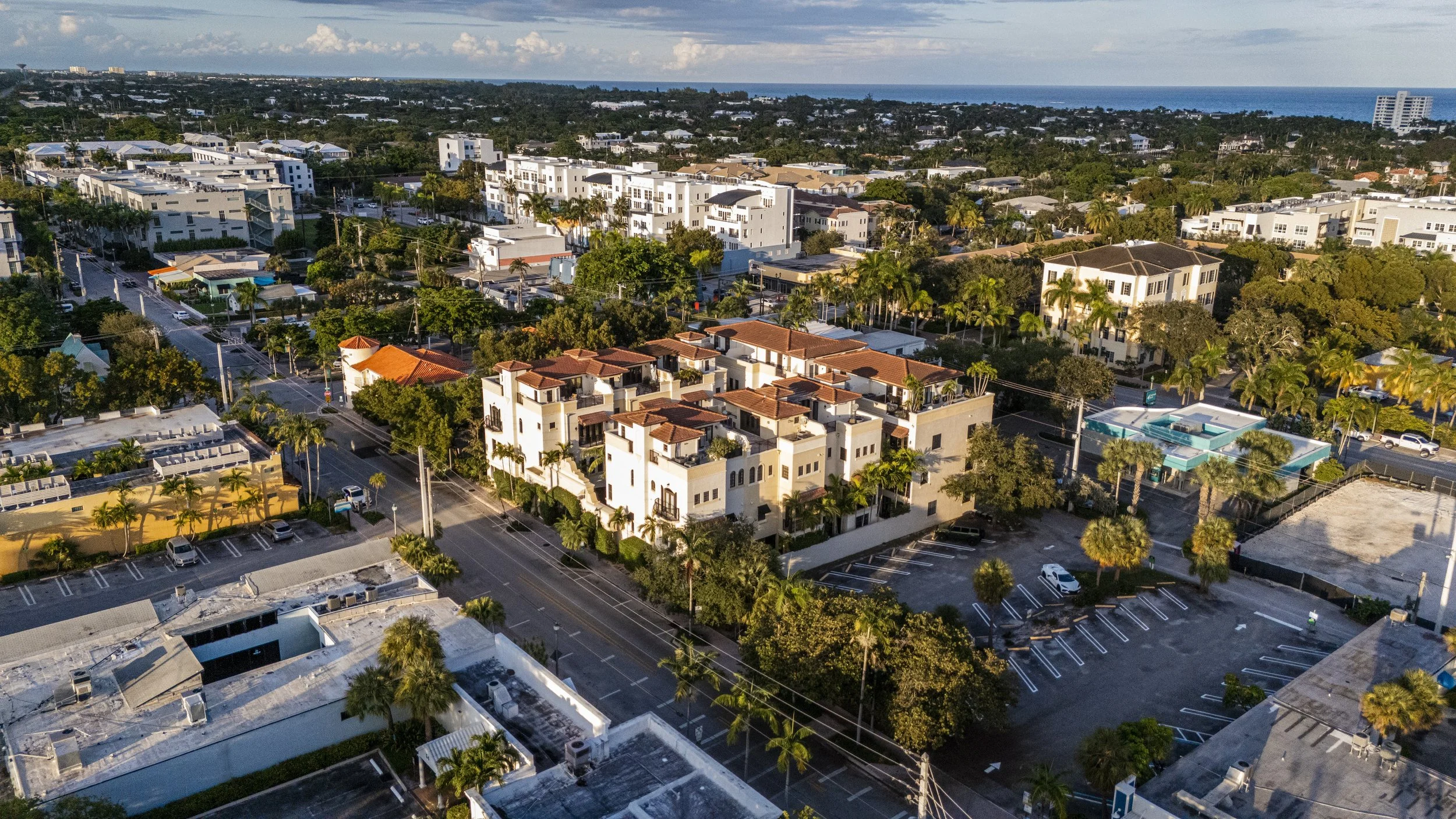 An aerial view of a city neighborhood with various residential buildings, streets, trees, and a distant ocean view.