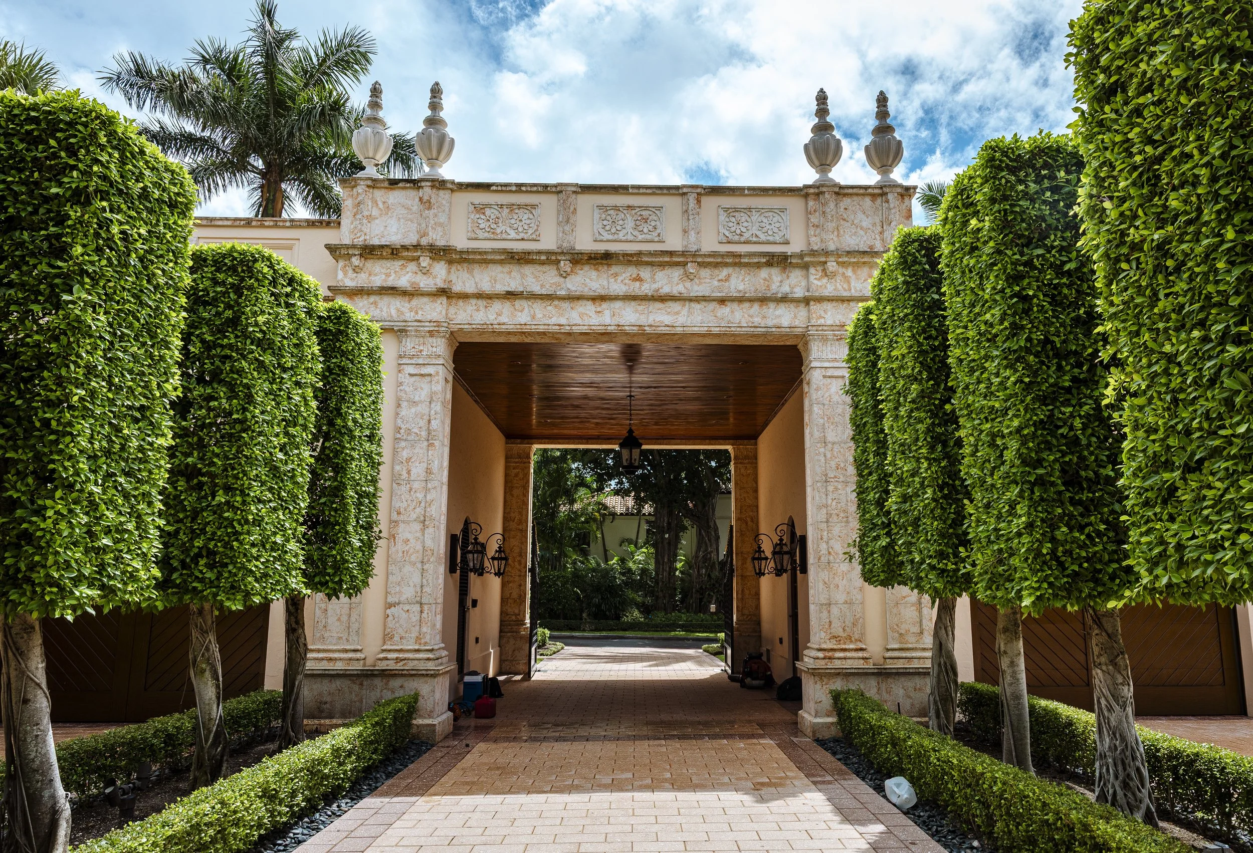 Entrance to a luxurious residence with stone archway, tall manicured trees lining the walkway, and a wooden ceiling under the arch.