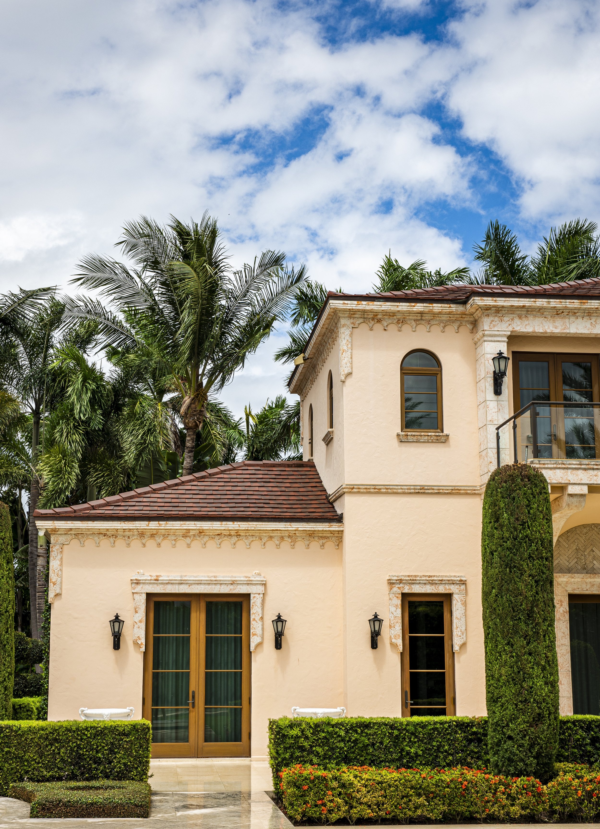 A large, beige, stucco house with a red tile roof, surrounded by lush tropical trees and well-manicured bushes, under a partly cloudy blue sky.
