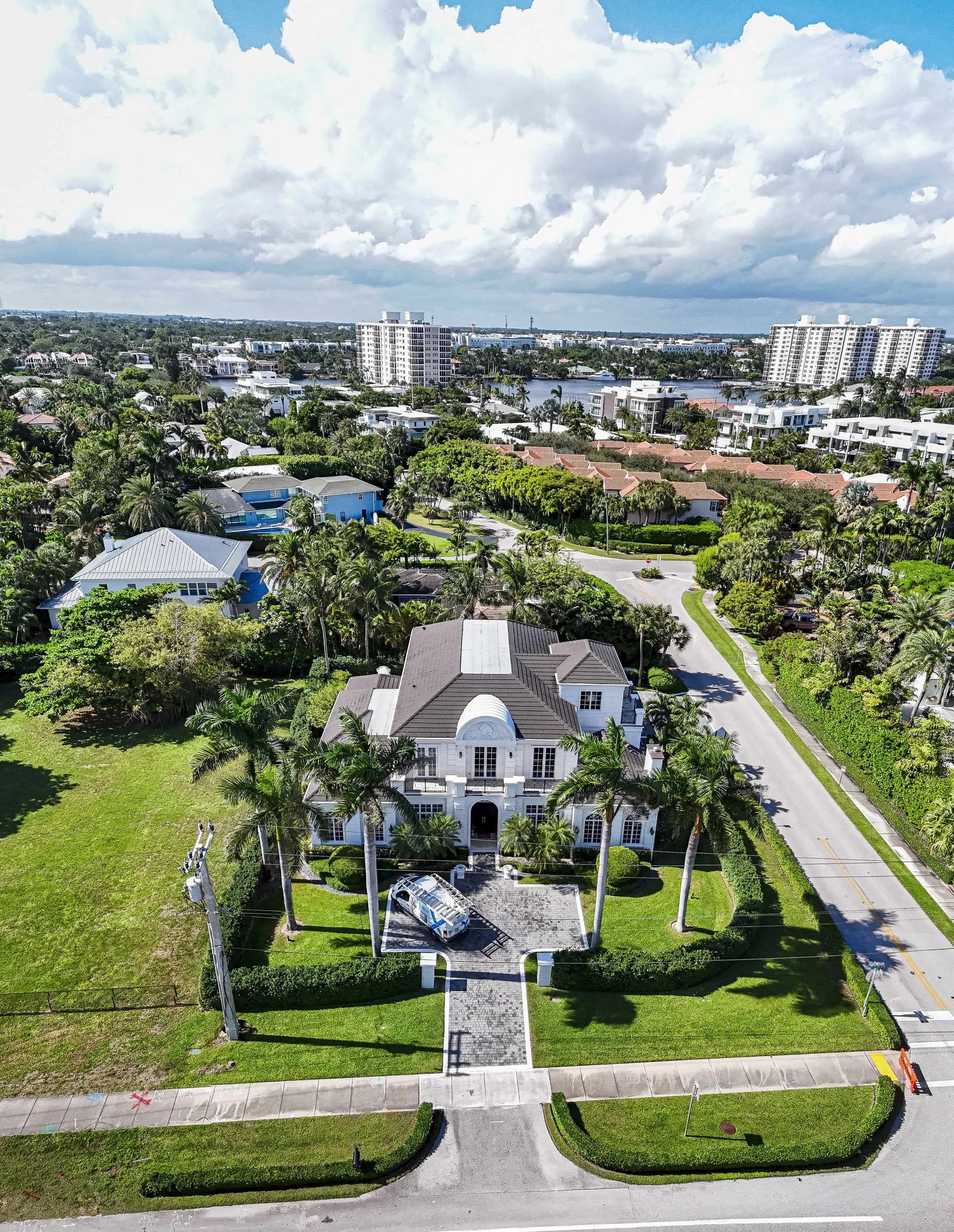 Aerial view of a large, white, two-story house with a curved facade, surrounded by tall palm trees and green lawn, located in a suburban neighborhood with other houses and an apartment complex nearby, under a partly cloudy sky.