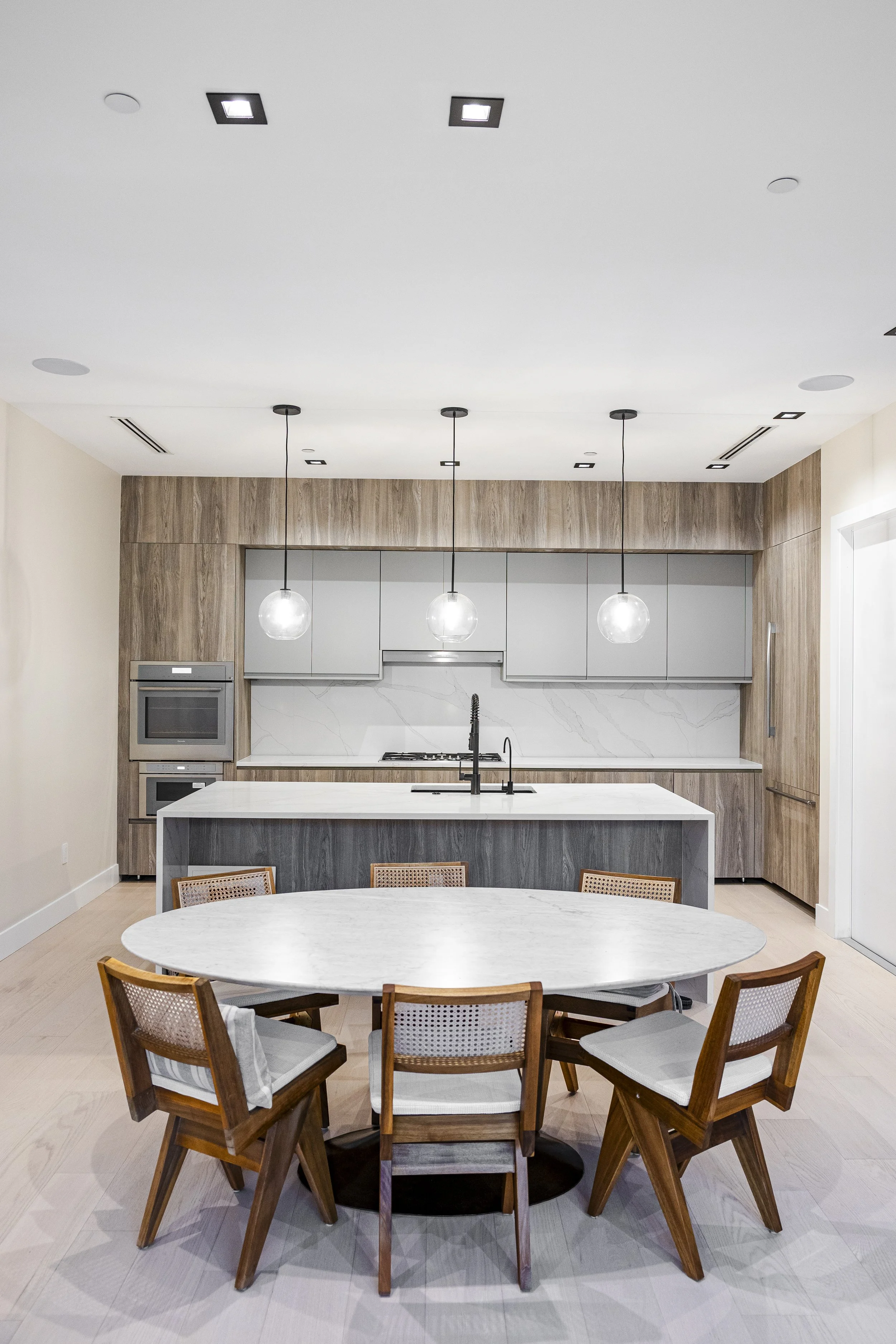 Modern kitchen with a white marble island, gray cabinets, wooden wall panels, and hanging spherical pendant lights, with a round dining table and six wooden chairs.
