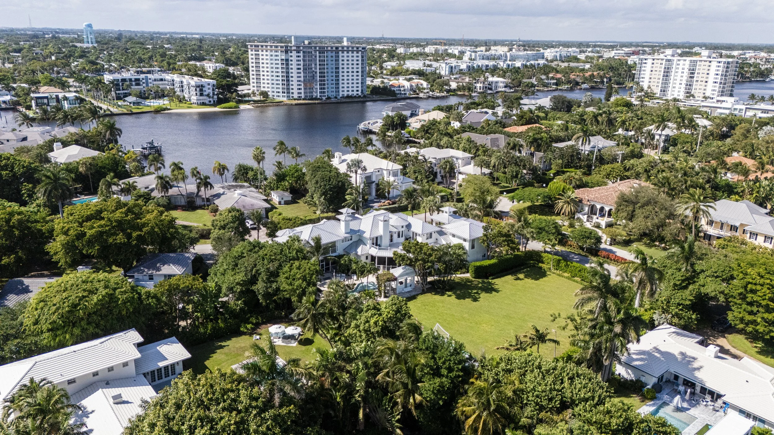 Aerial view of a lush residential area with houses, trees, and a waterway, with tall buildings in the background under a partly cloudy sky.