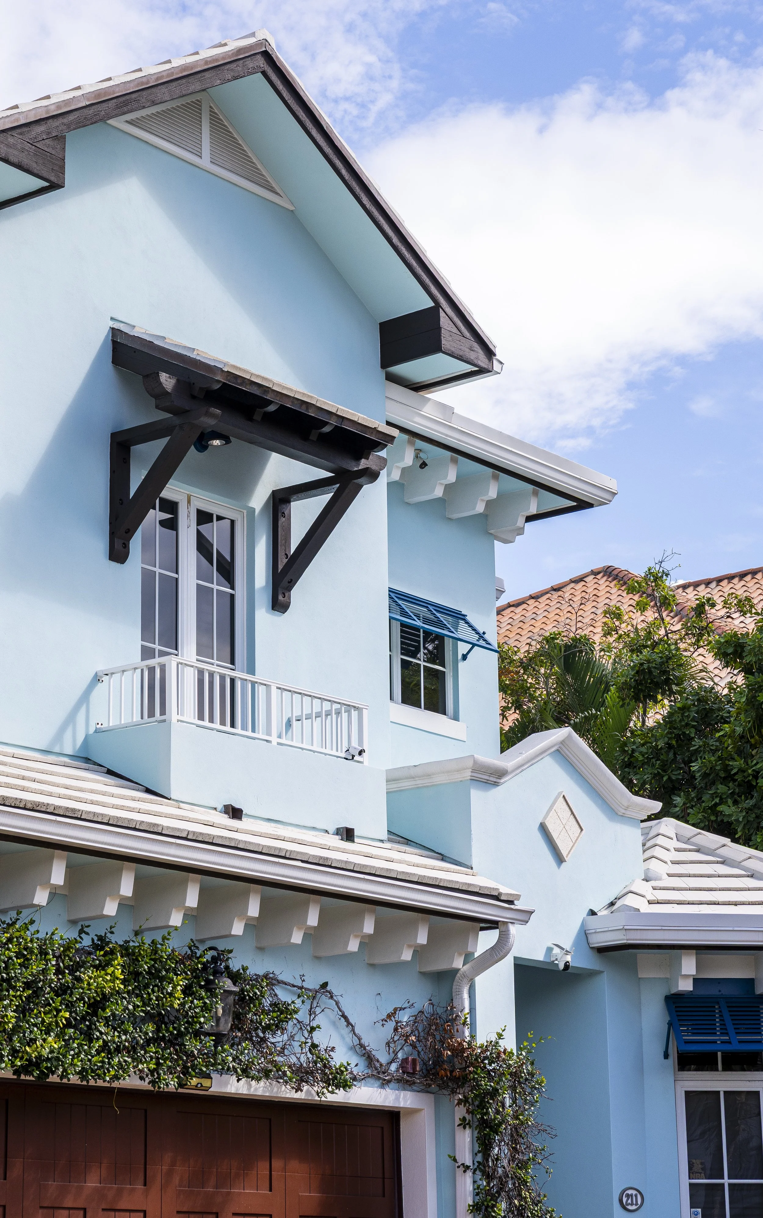 Bright blue multi-story house with white trim and black accents, featuring a small balcony, multiple windows with blue shutters, and a sloped roof with tiled roofing. There are bushes and vines near the lower part of the house, under a partly cloudy 