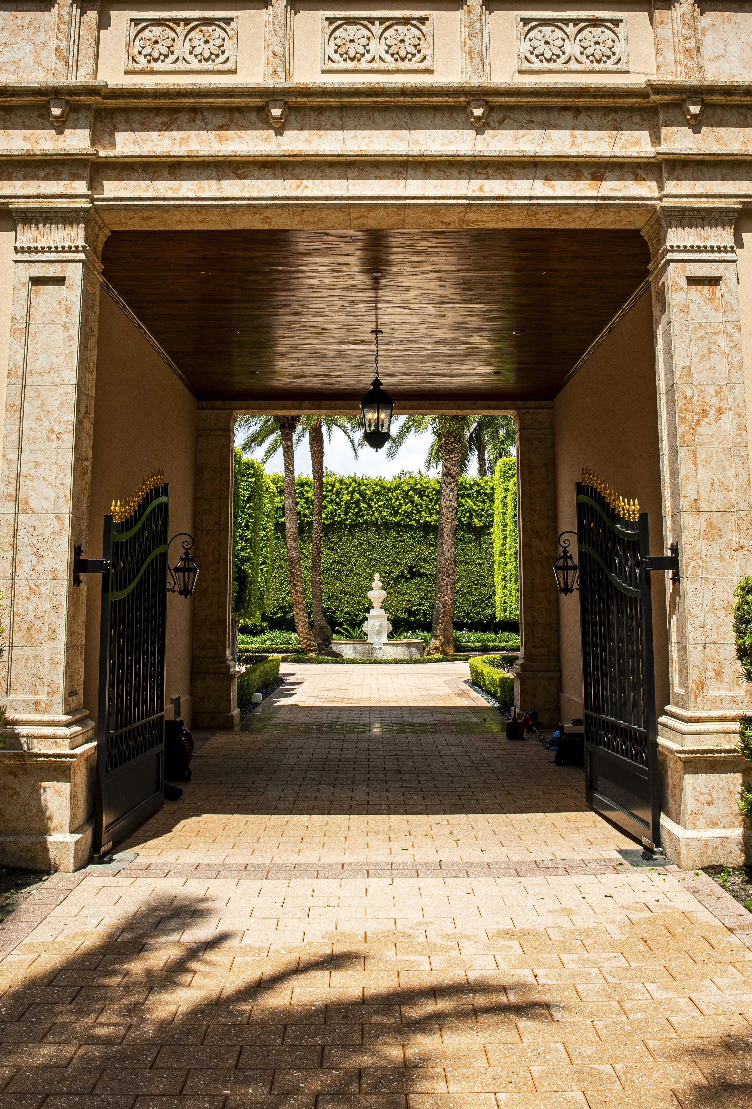 Open wrought iron gates leading to a courtyard with a stone fountain and neatly trimmed hedges, surrounded by tall palm trees and lush greenery.