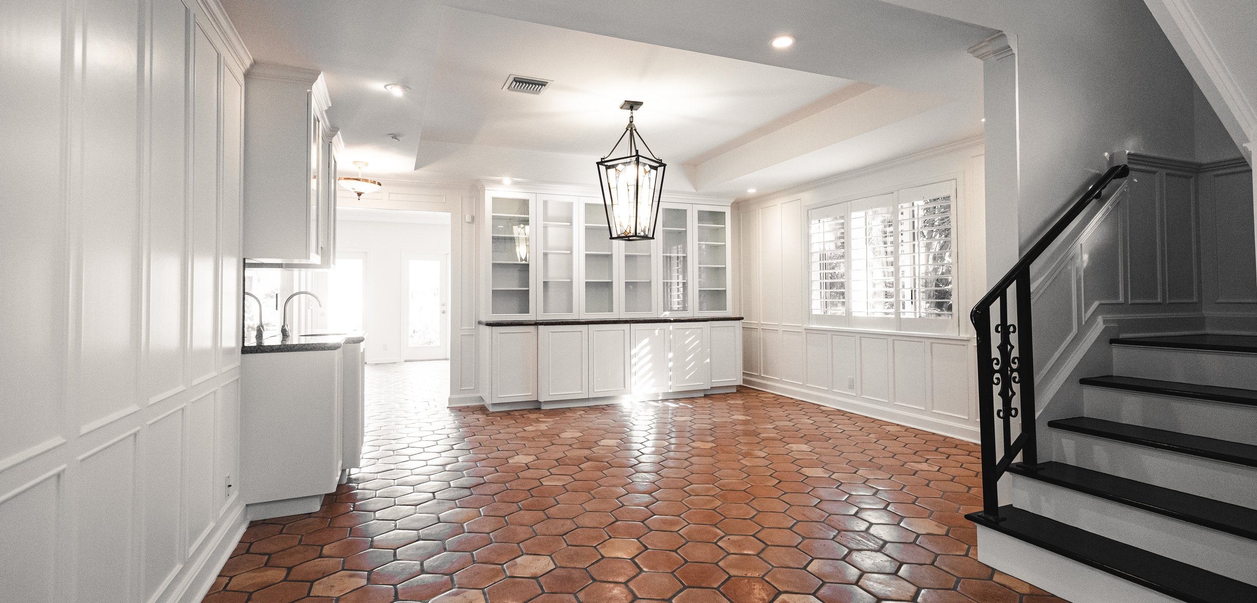 Bright, spacious room with white paneled walls, terracotta hexagonal tile floor, large windows with plantation shutters, and a black stair railing.
