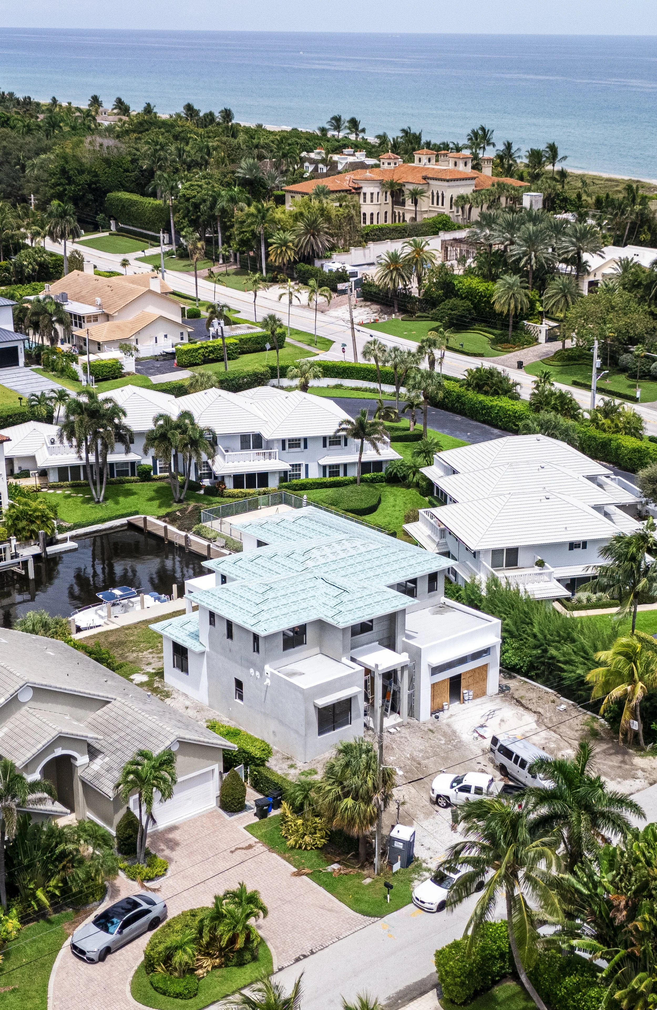 Aerial view of a coastal neighborhood with new construction homes, surrounded by palm trees and lush greenery, with the ocean in the background.