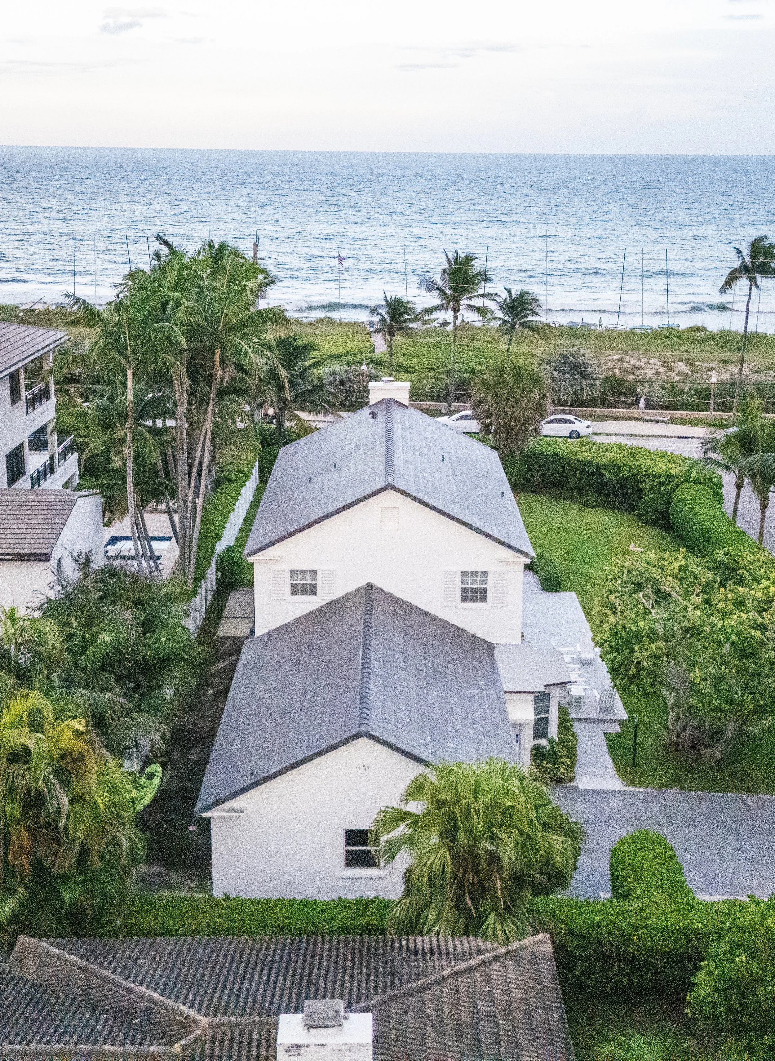 Aerial view of a house near the ocean with palm trees, a grassy yard, and neighboring buildings, overlooking the beach and water.