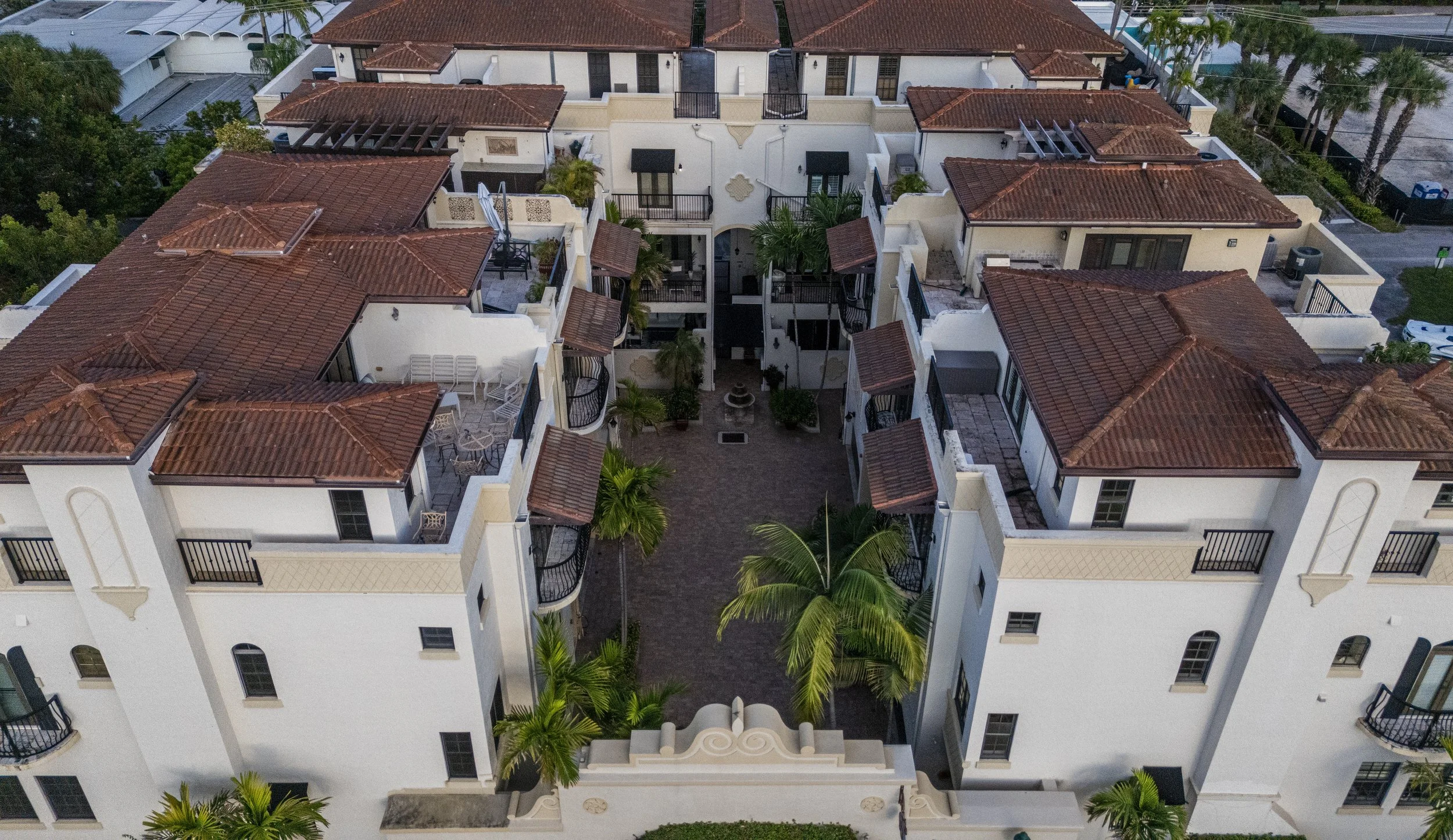 Aerial view of a Mediterranean-style apartment complex with white walls, red-tiled roofs, balconies, palm trees, walkways, and outdoor seating areas.
