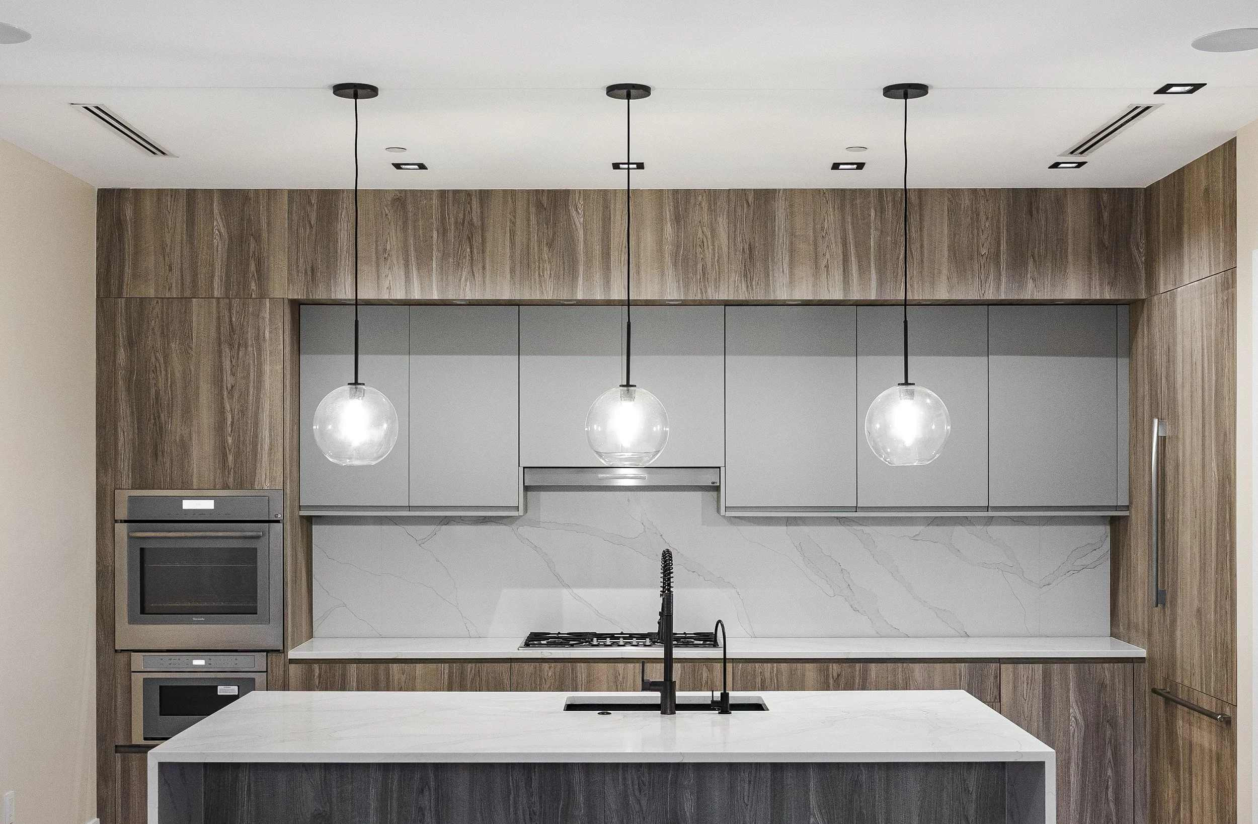 Modern kitchen with gray cabinets, marble backsplash, wooden wall panels, black faucet, and three glass pendant lights