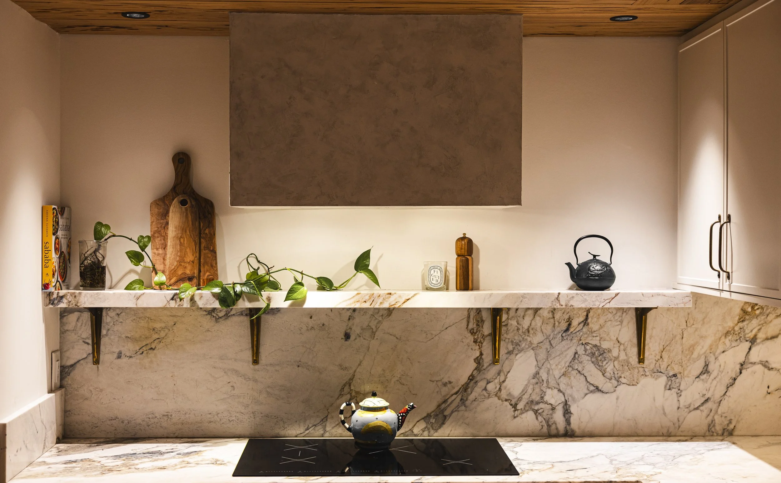 A kitchen countertop with decorative items, a plant, and a teapot on a marble surface, with cabinets on the right and a wooden tray and books on the left side.