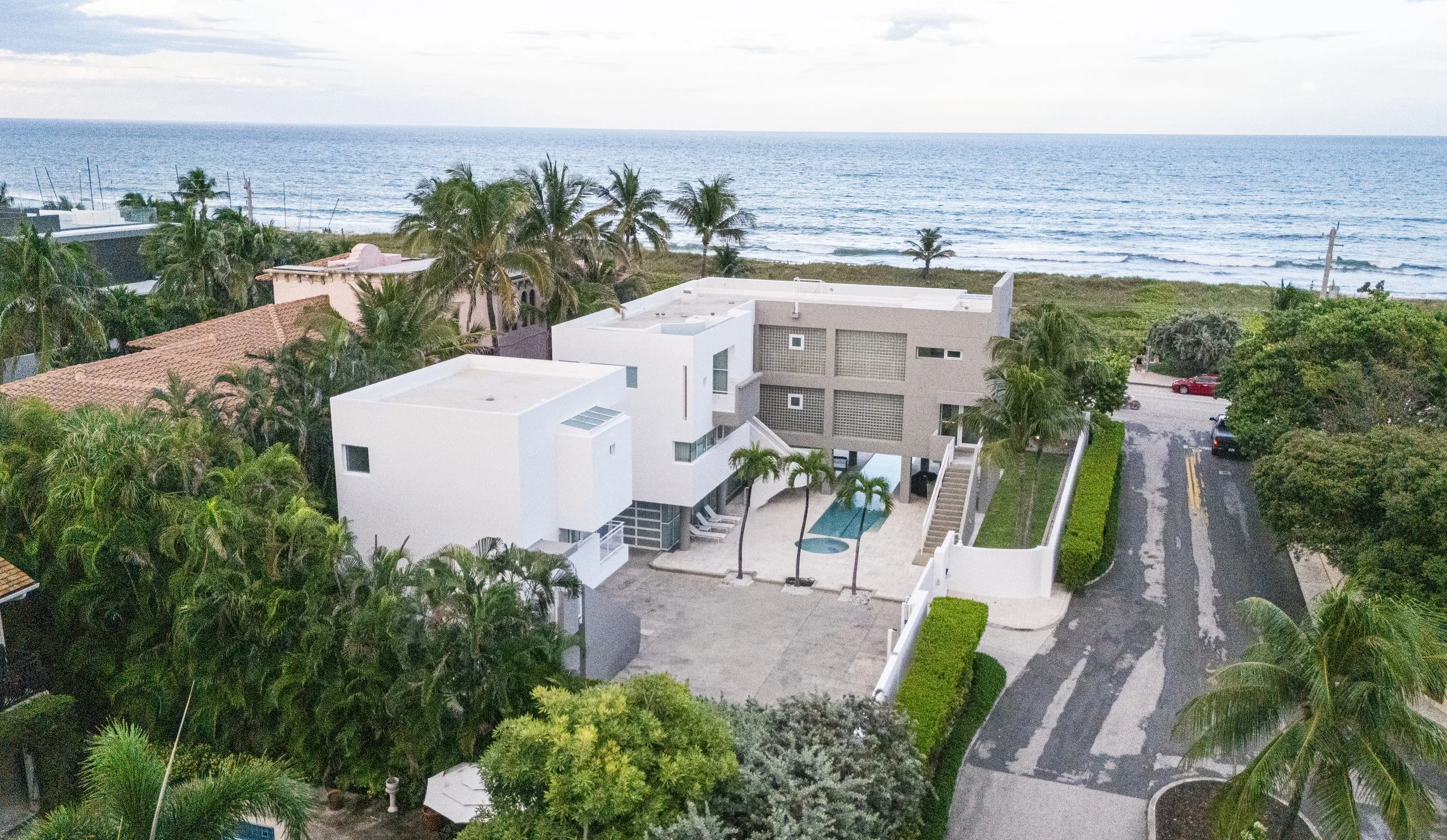 Aerial view of a modern white beach house with a small swimming pool, surrounded by palm trees, neighboring houses, and a road leading to the ocean.